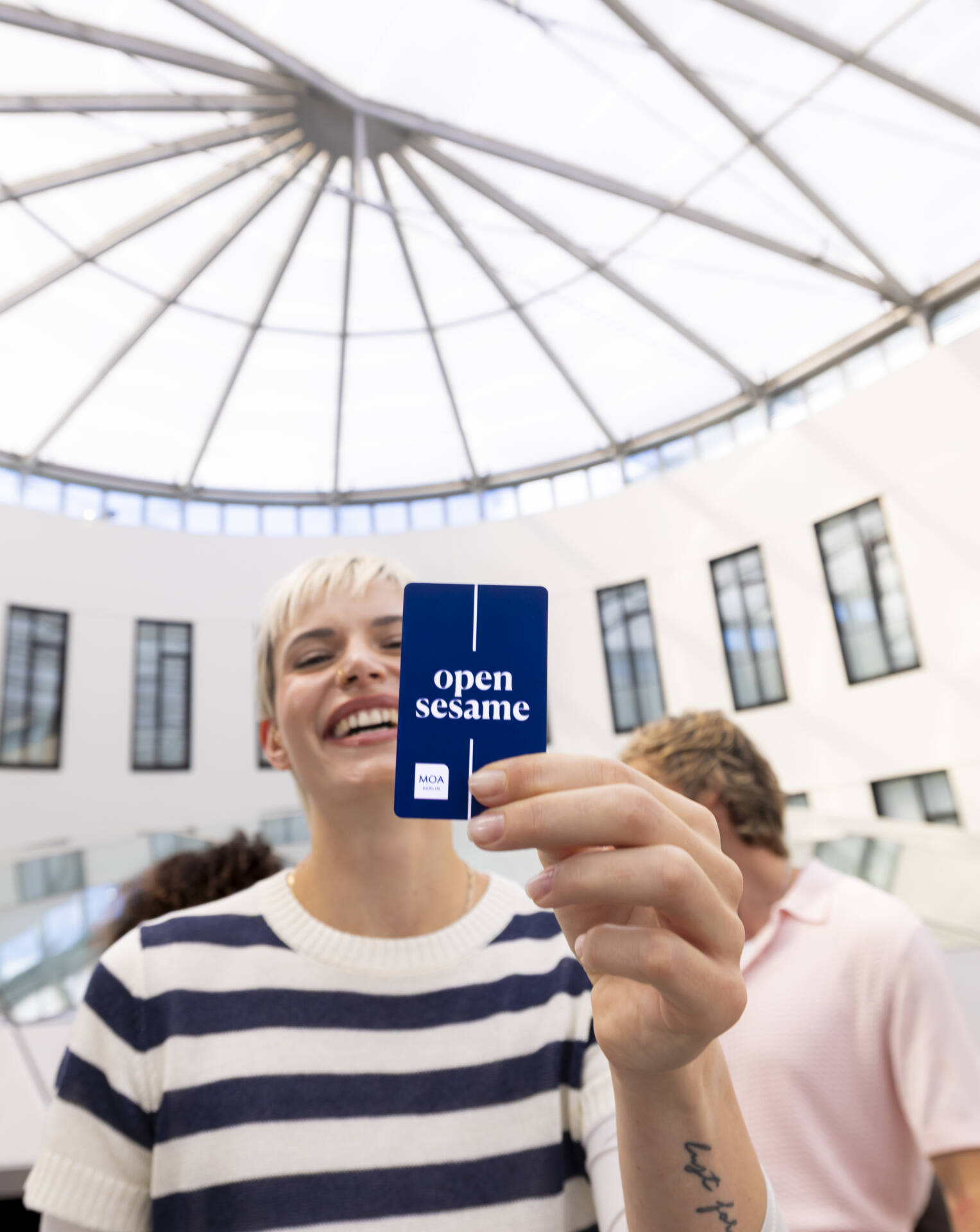 Eine Person hält eine blaue Karte mit dem weißen Schriftzug „open sesame“ in einem hellen Atrium mit Glasdach in die Kamera.