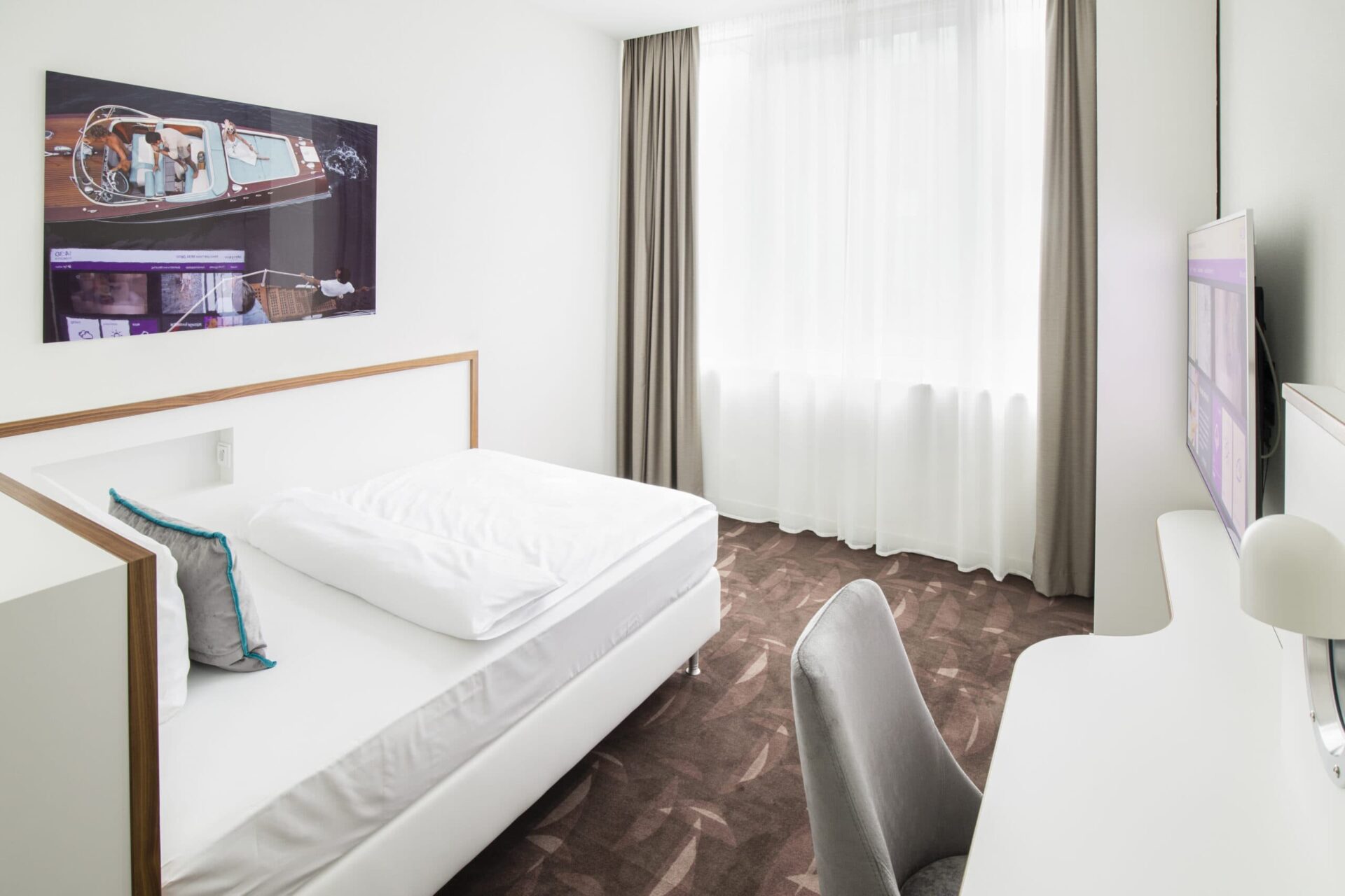 Ein schlicht eingerichtetes Hotelzimmer in Berlin mit einem Einzelbett, Schreibtisch, Stuhl, einer großen Fensterfront mit Vorhängen und einem Bild an der Wand.