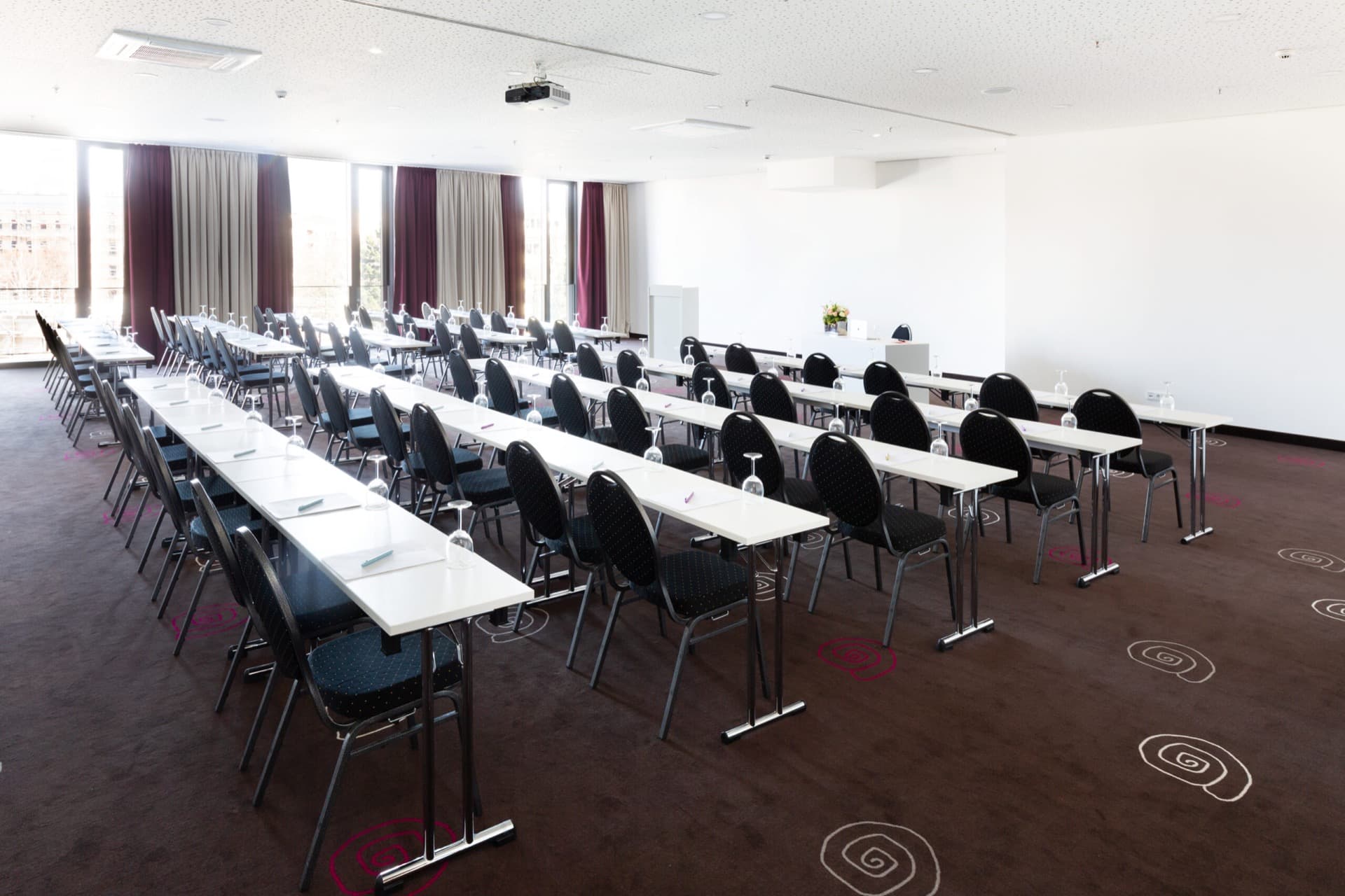 A bright seminar room with parliamentary row seating, tables, chairs, a screen and windows.