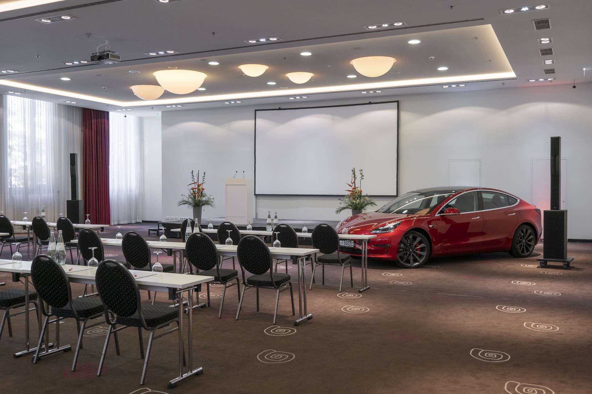 Ein roter Tesla mit geöffneter Fahrertür in großem, leerem Konferenzraum mit langen Tischen und Stühlen im Hotel MOA Berlin.