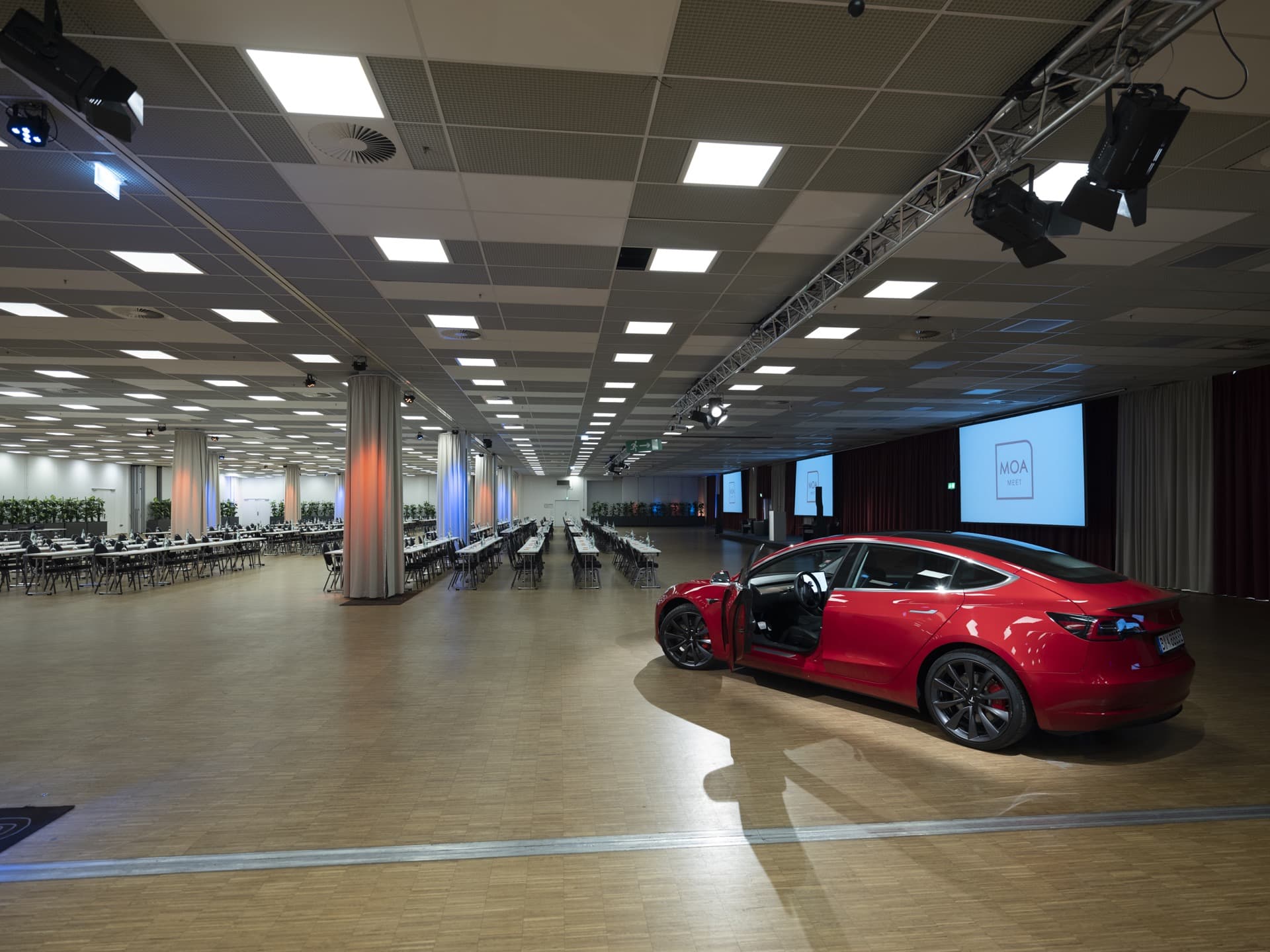 A red Tesla with the driver's door open in a large, empty conference room with long tables and chairs at Hotel MOA Berlin.