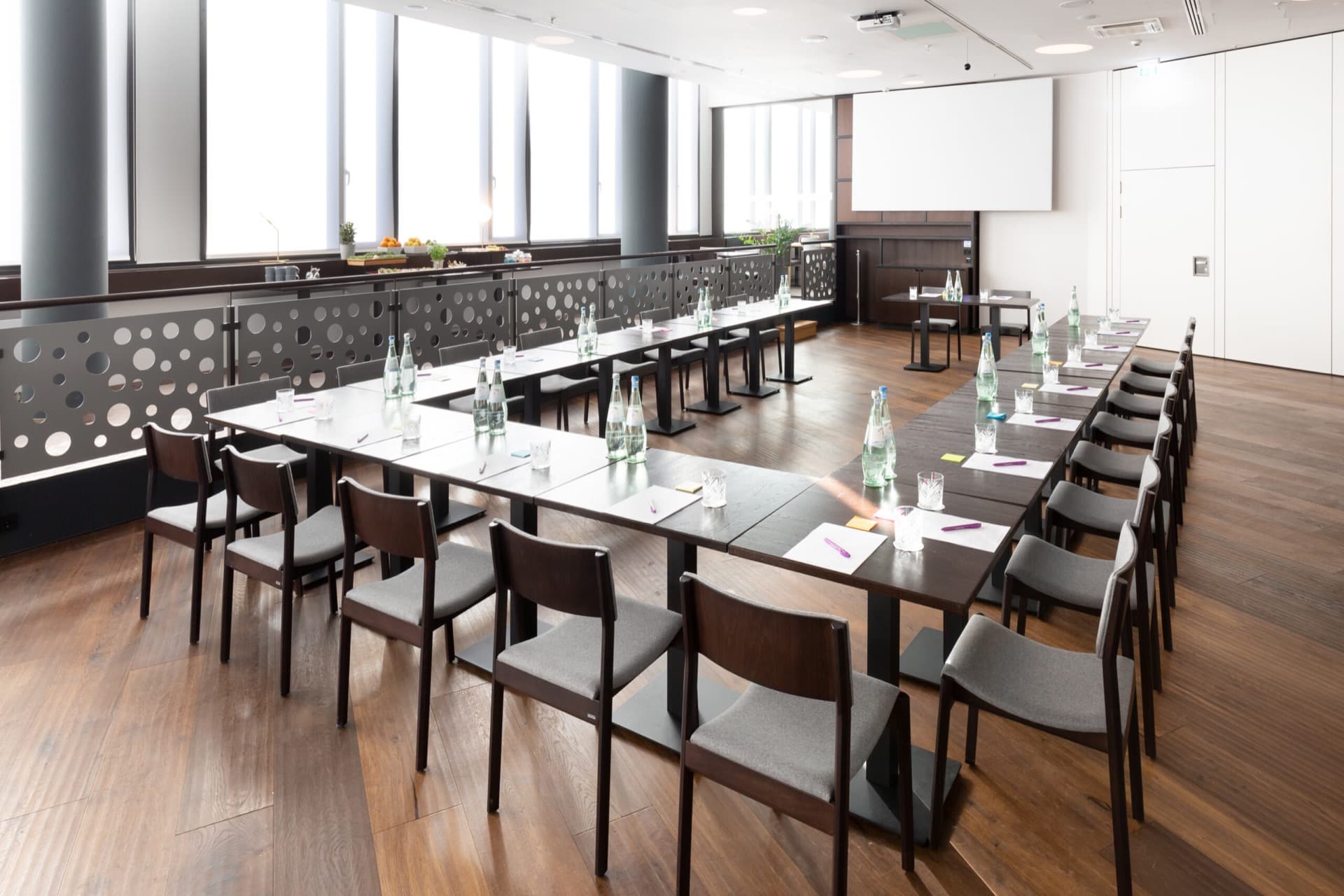 Ein moderner Konferenzraum mit einer parlamentarischen Bestuhlung, Tischen, Stühlen, einer modernen Präsentationstechnik und Tageslicht für Tagungen.