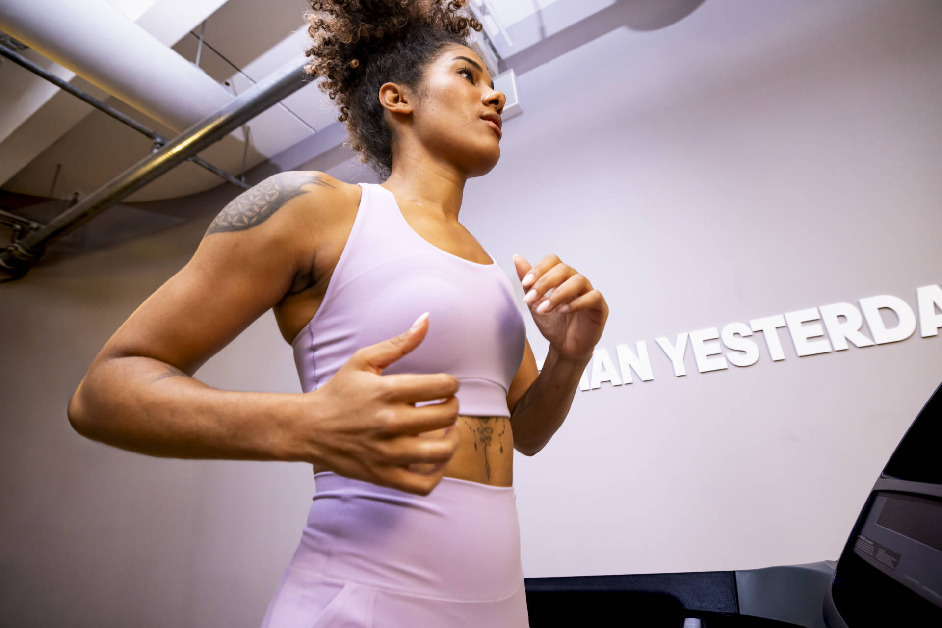 Eine Frau in lila Sportkleidung läuft auf einem Laufband in einem Fitnessraum mit sichtbaren Rohrleitungen und Wandbeschriftung.