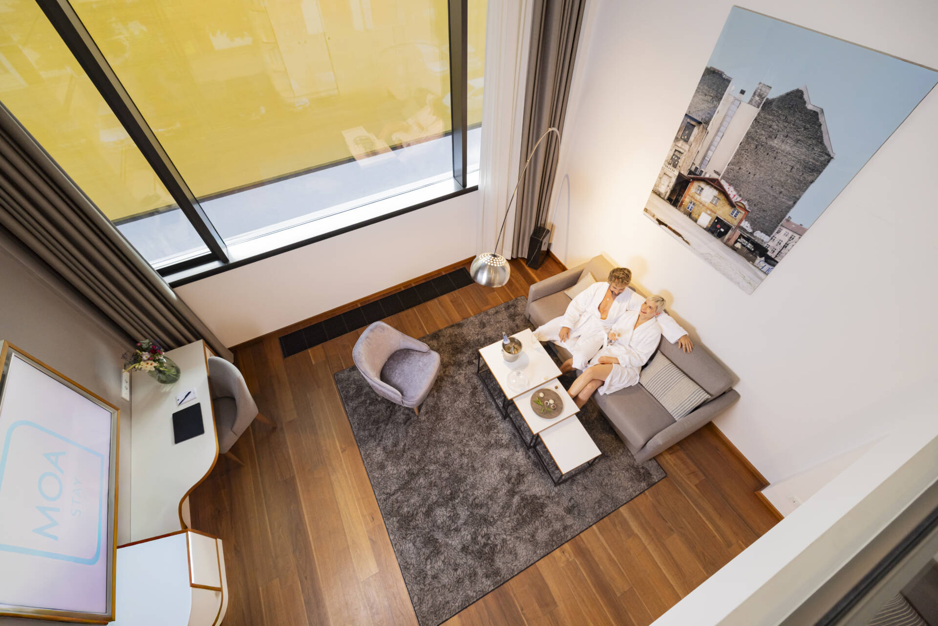 Zwei Personen sitzen auf einem Sofa in einem modernen Wohnbereich mit Couchtisch, Teppich, Sessel, Schreibtisch und großem Oberlicht.