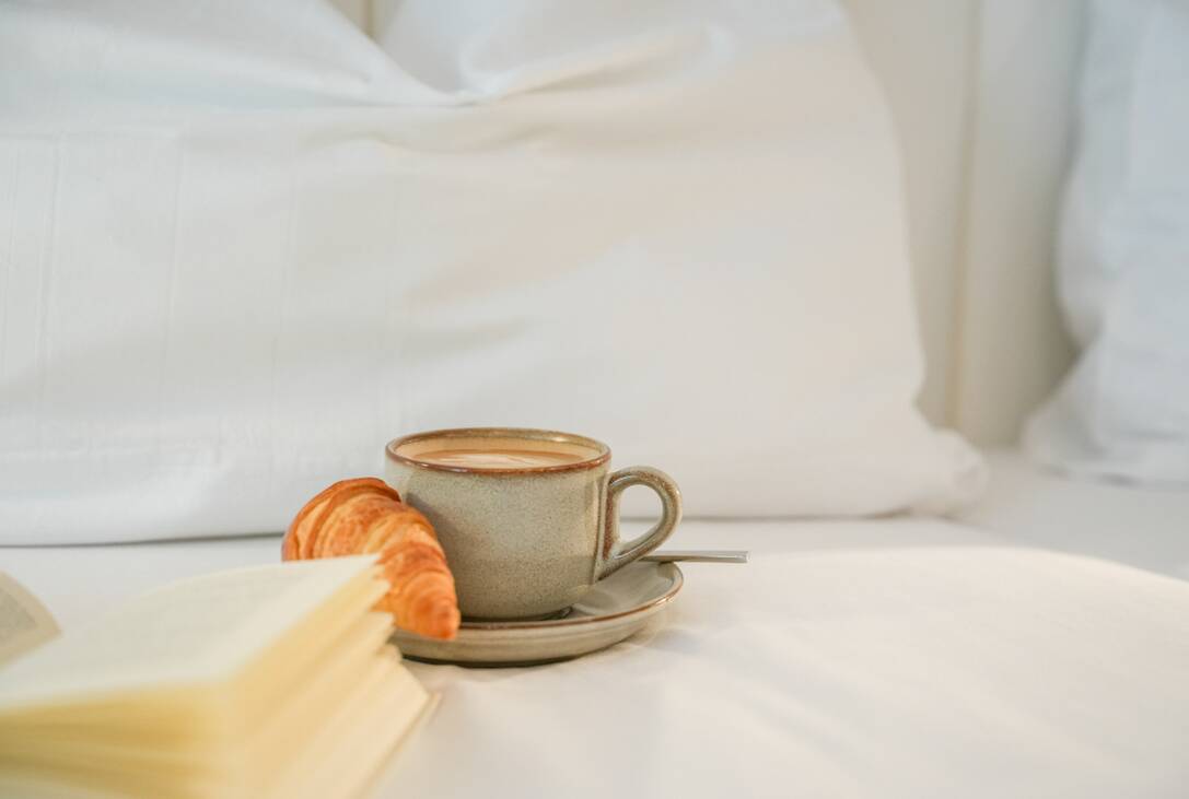 Eine Tasse Kaffee mit Untertasse und Croissant steht neben einem aufgeschlagenen Buch auf einem weißen Bett.