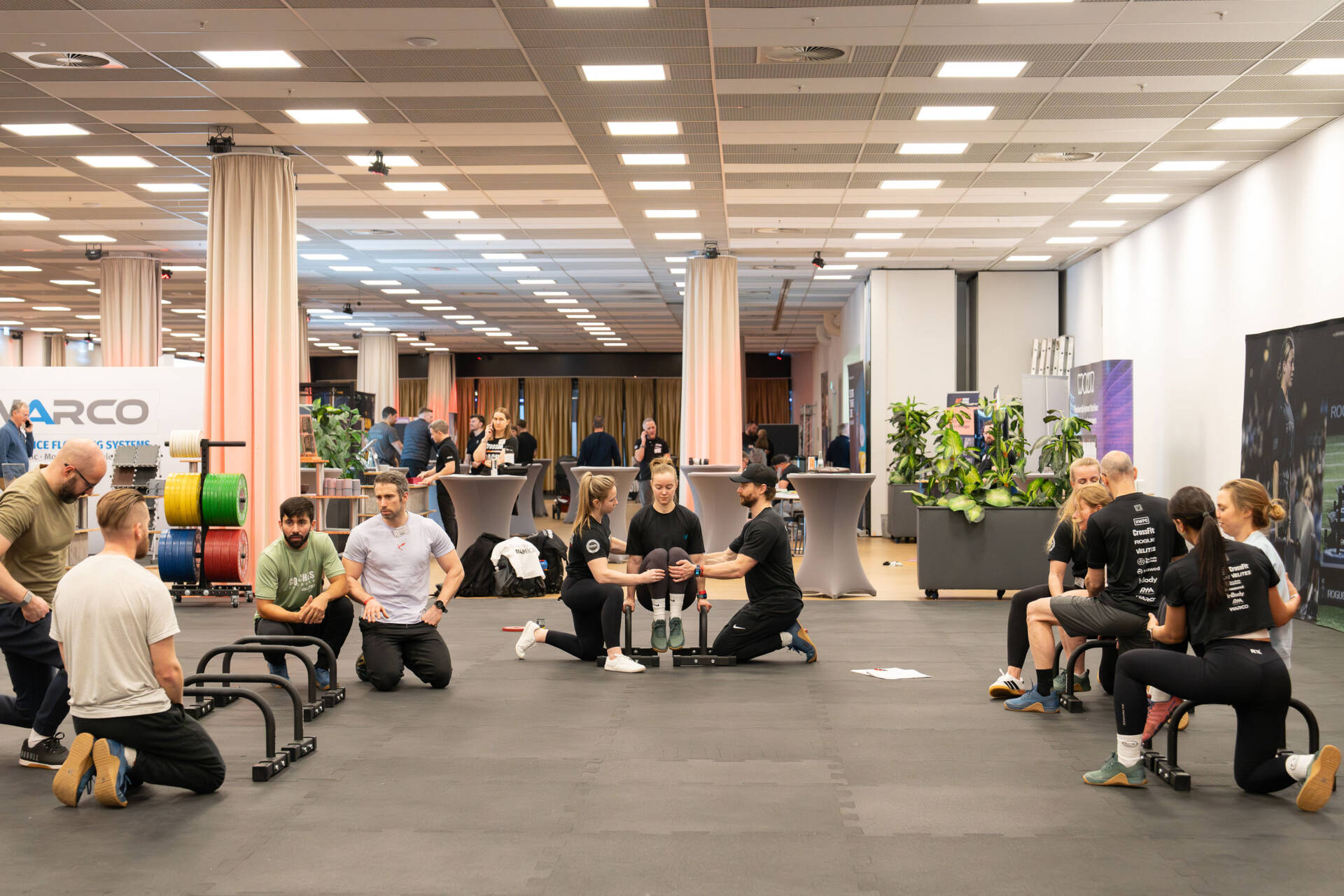 Menschen trainieren in einem hellen Fitnessbereich in Gruppen mit Hanteln, Matten und weiteren Trainingsgeräten.