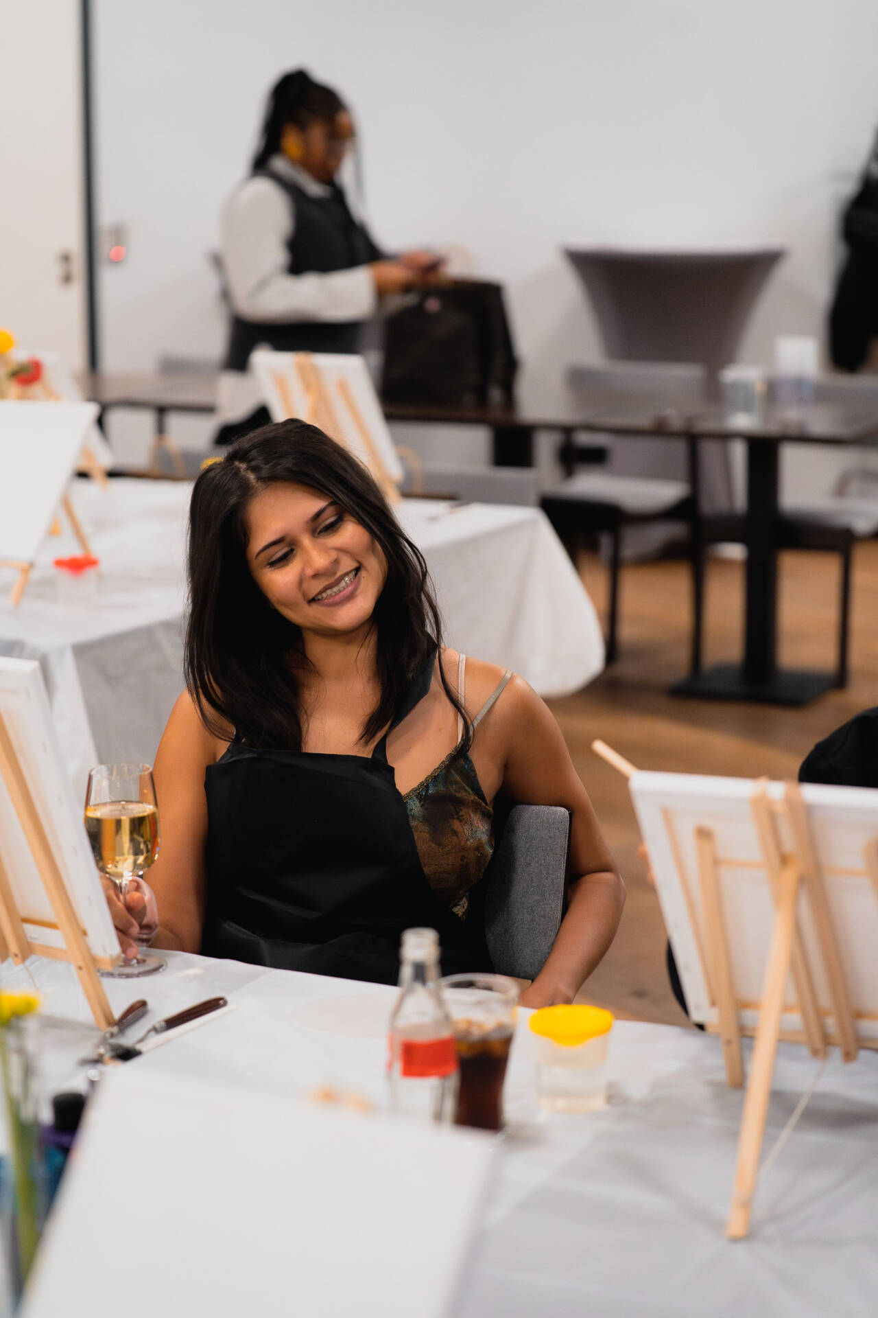 A woman sits at a set table at an event and holds a glass of sparkling wine, while more tables stand in the room.