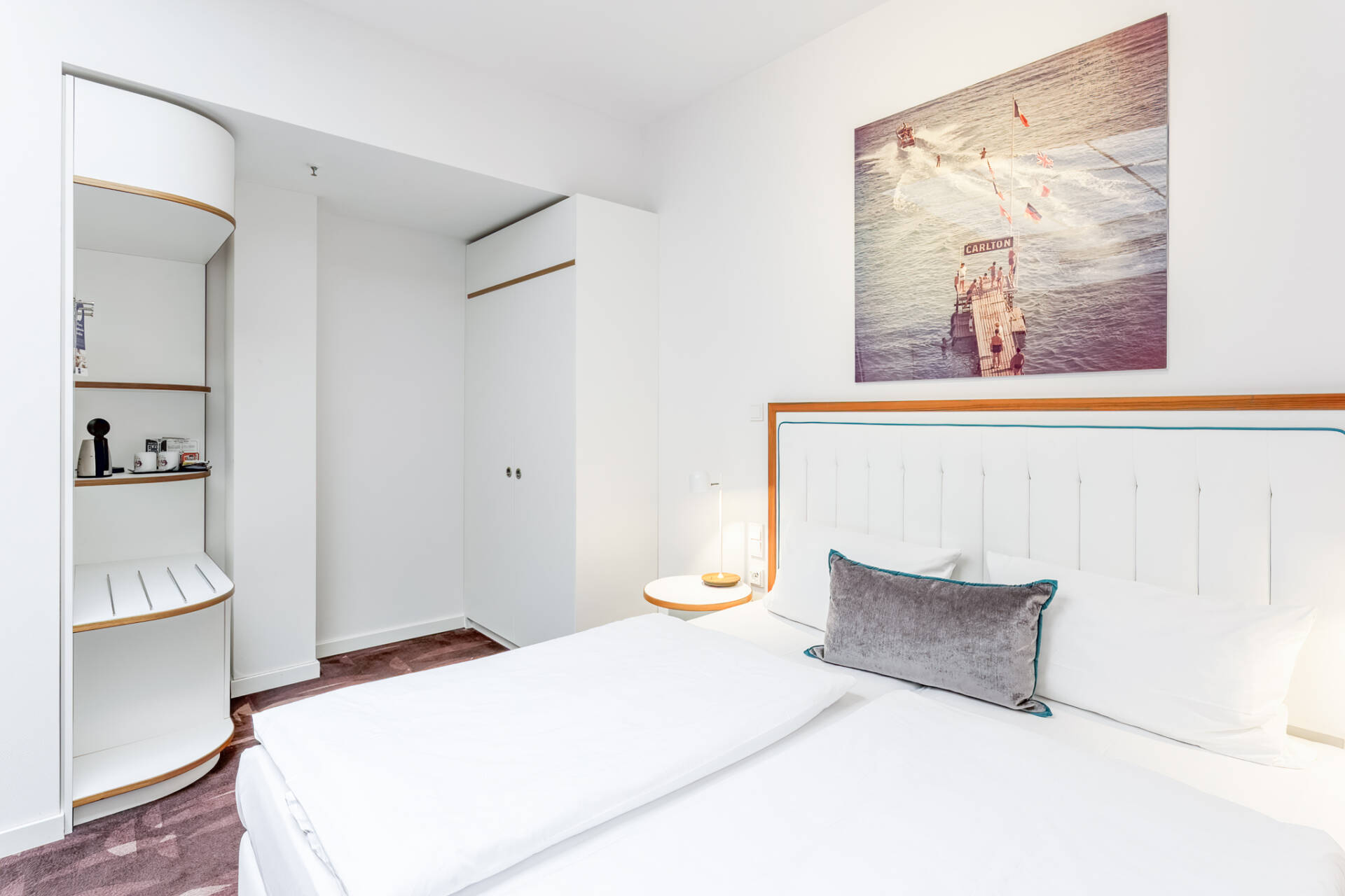 Helles Hotelzimmer mit einem Doppelbett, integrierten weißen Regalen, Nachttischen, einer Tischlampe und einem gerahmten Bild an der Wand.