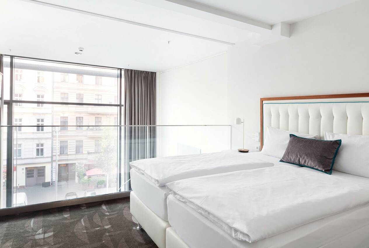 Bright bedroom with a large double bed, white headboard, floor-to-ceiling windows, curtains and a view of surrounding buildings.