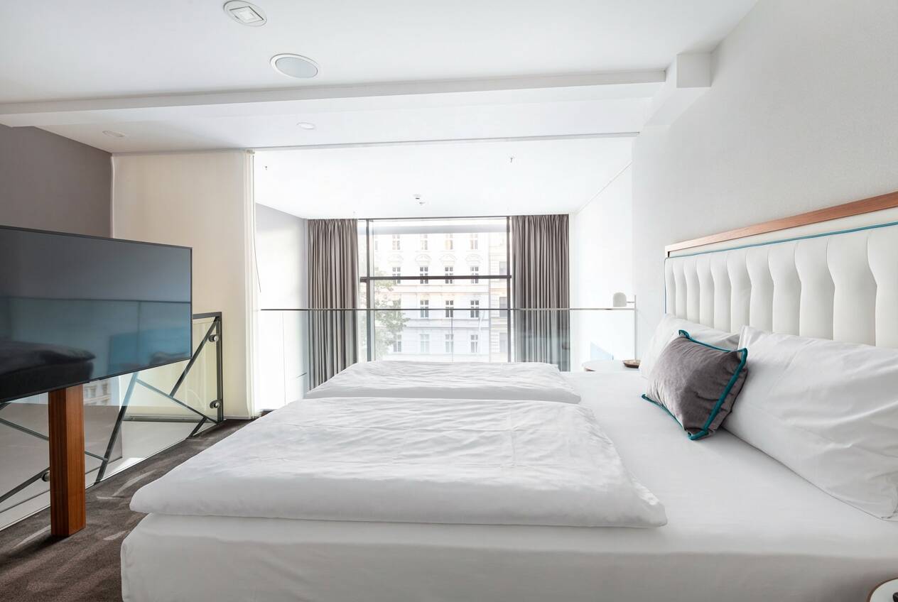 Helles Hotelzimmer mit großem Doppelbett, gepolstertem Kopfteil, Fernseher, Vorhängen und bodentiefem Fenster.