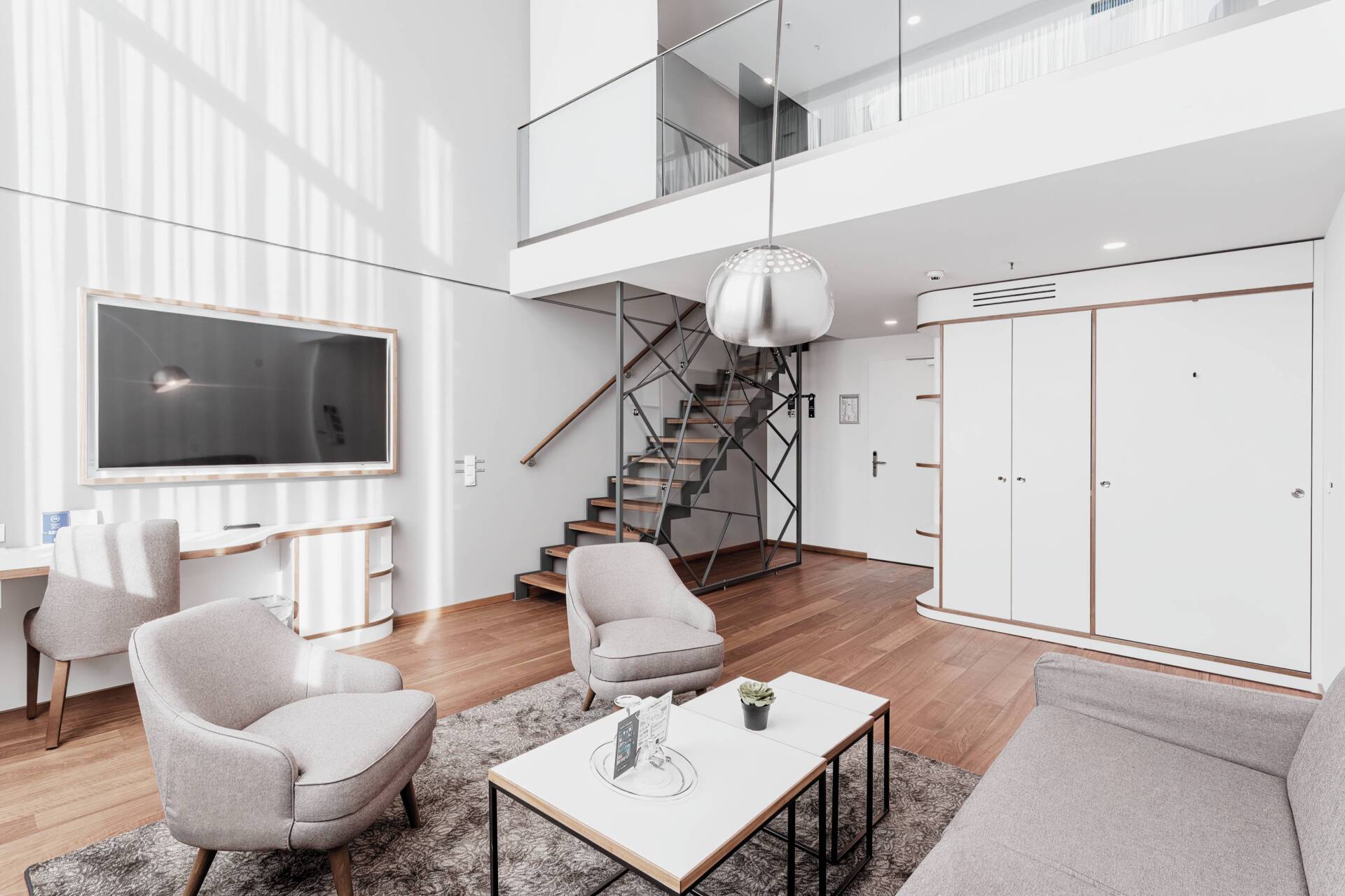 Heller Sitzbereich einer Hotelsuite mit zwei Sesseln, Sofa, Couchtisch, Fernseher, Holztreppe und Galerie.