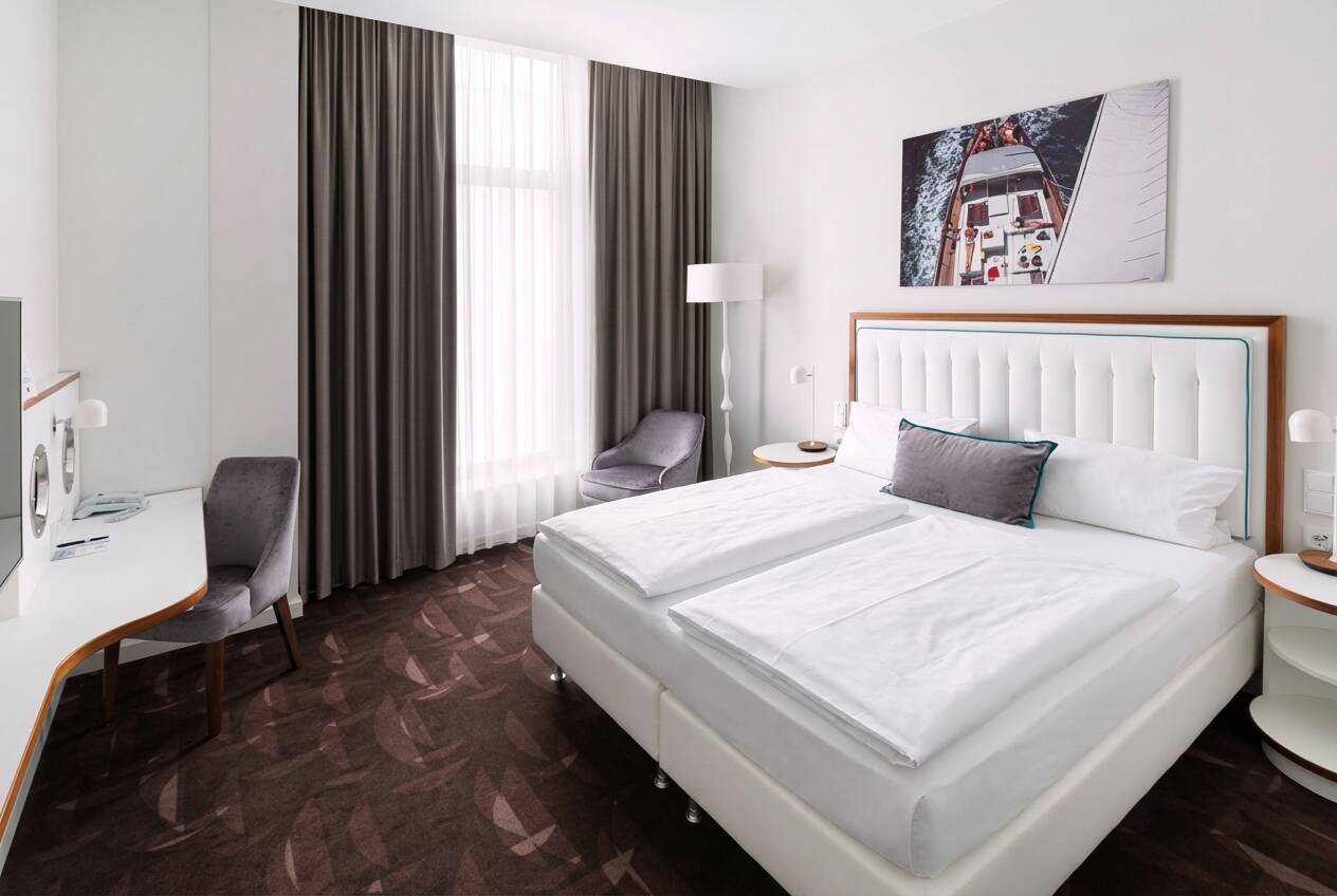 Modern eingerichtetes Hotelzimmer mit Doppelbett, Schreibtisch, Sesseln, großen Fenstern mit Vorhängen und Wandbild.