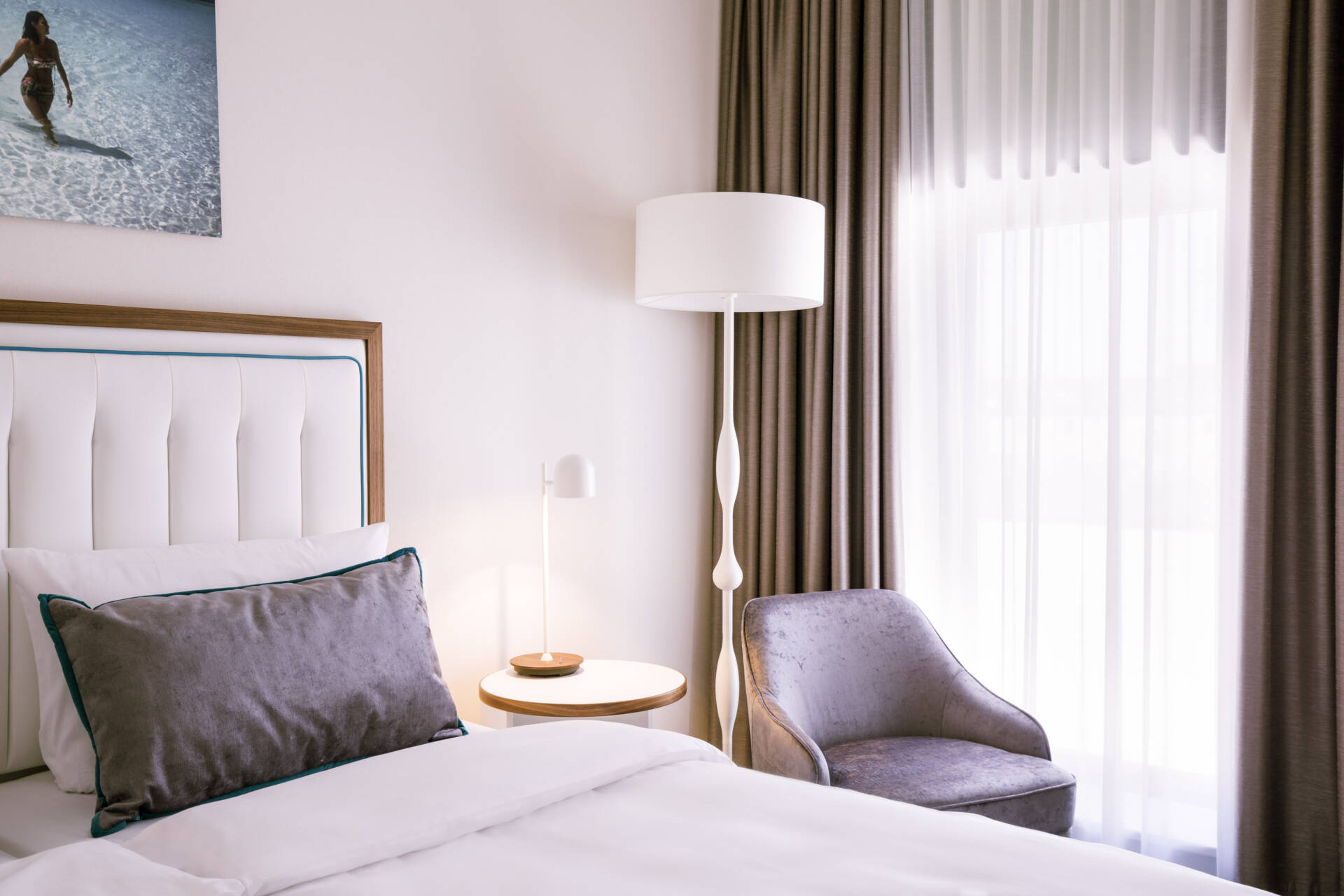 Modern eingerichtetes Hotelzimmer mit Doppelbett, Sessel, Beistelltisch, Stehlampe, Vorhängen und einem Bild an der Wand.