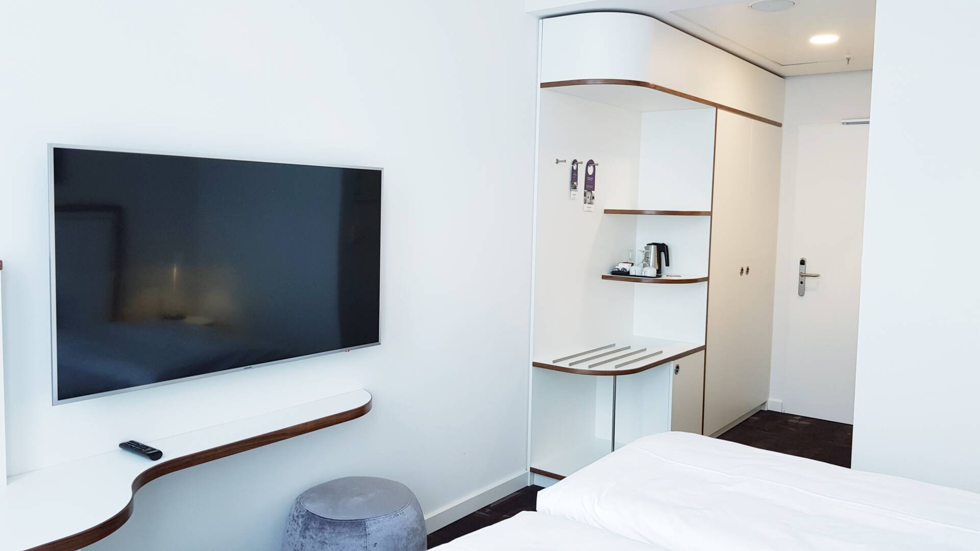 Helles Hotelzimmer mit großem Wandfernseher, integrierter Ablage, Sitzhocker, Kaffee- und Teestation sowie teilweise sichtbarem Bett.