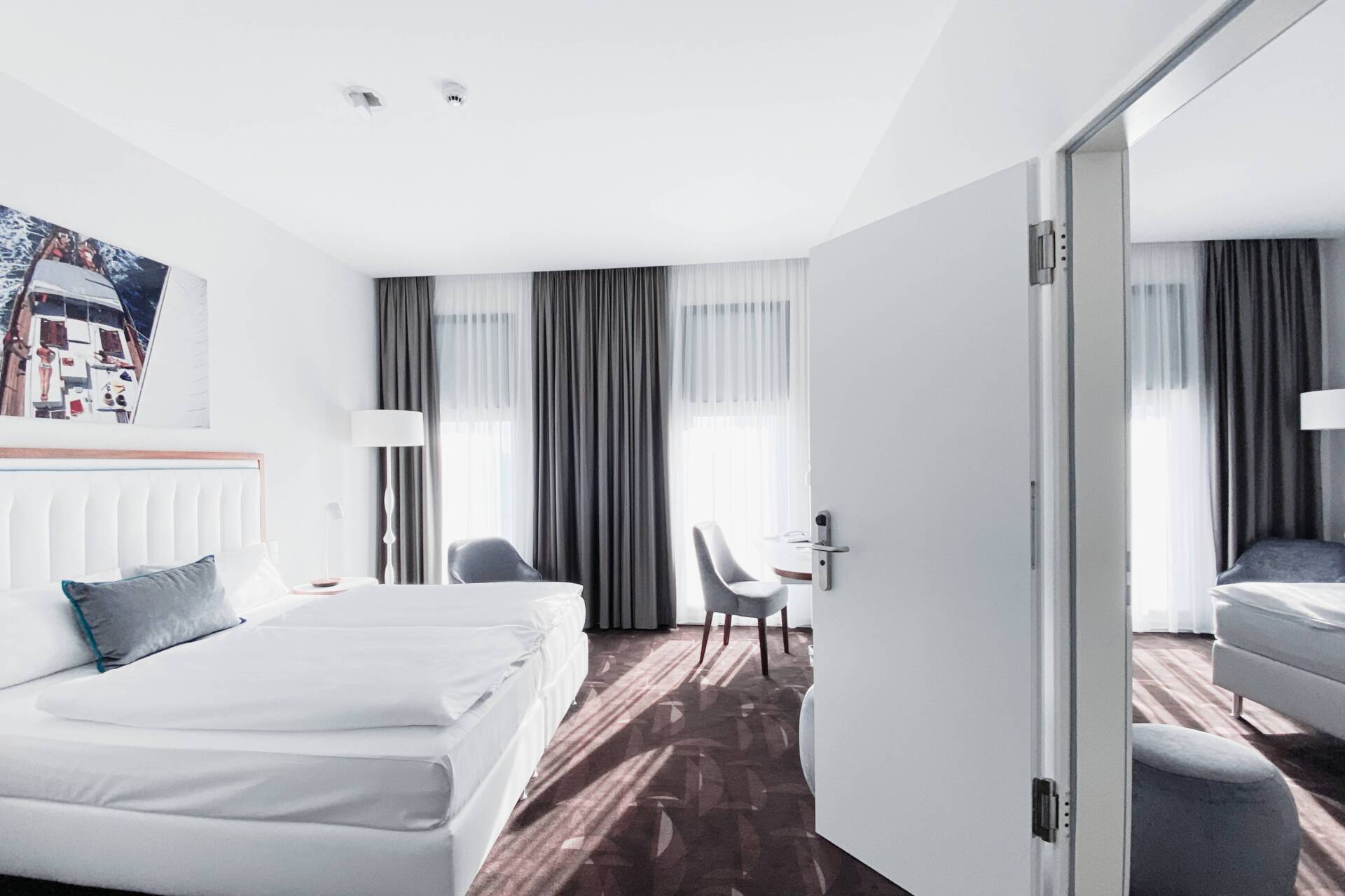 Helles Hotelzimmer mit Doppelbett, Sessel, Stuhl, Vorhängen, Wandbild und geöffneter Verbindungstür zu einem weiteren Zimmer.