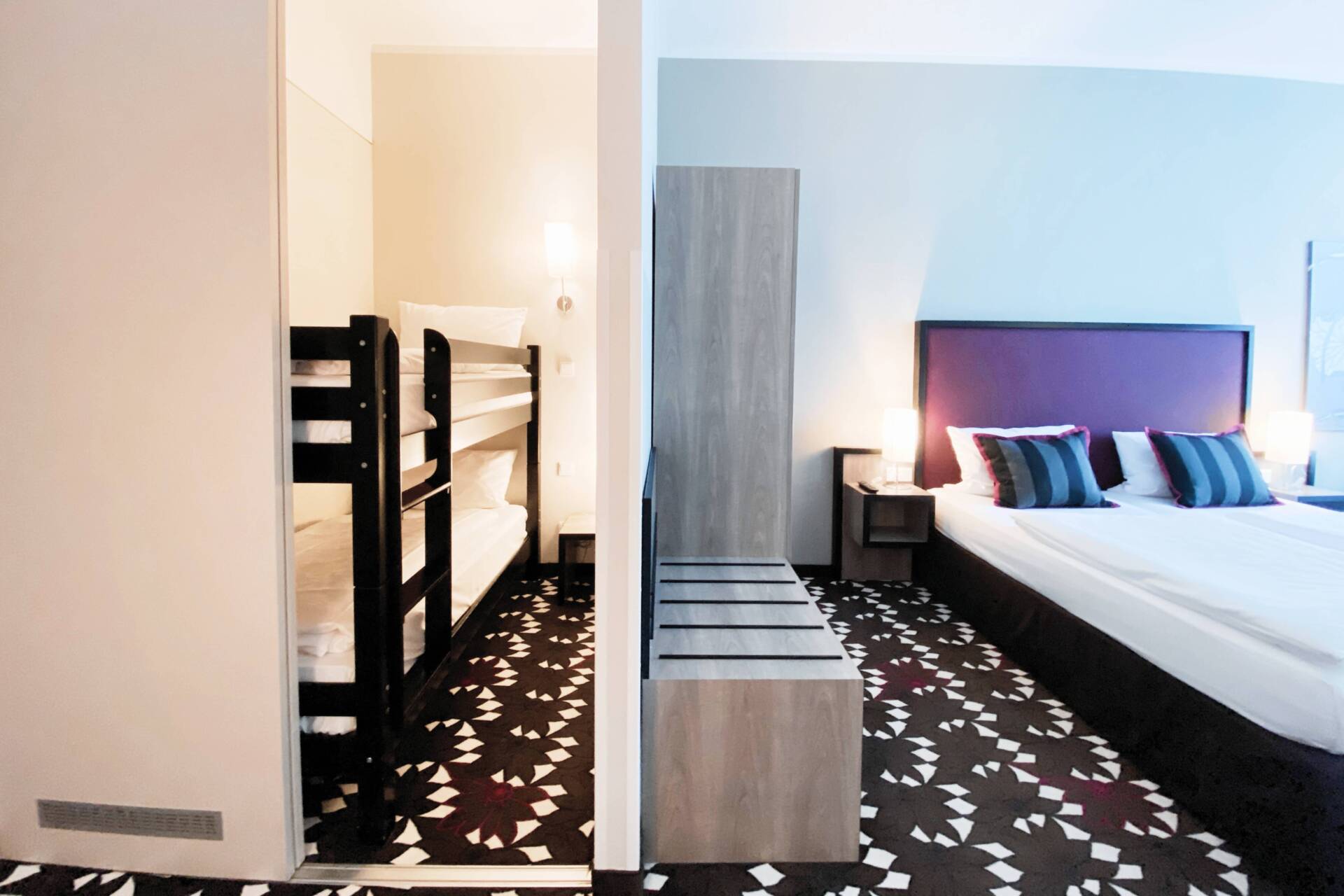 Helles Hotelzimmer mit Etagenbett, Doppelbett, Nachttischen, Schrank und schwarz-weiß gemustertem Teppichboden.