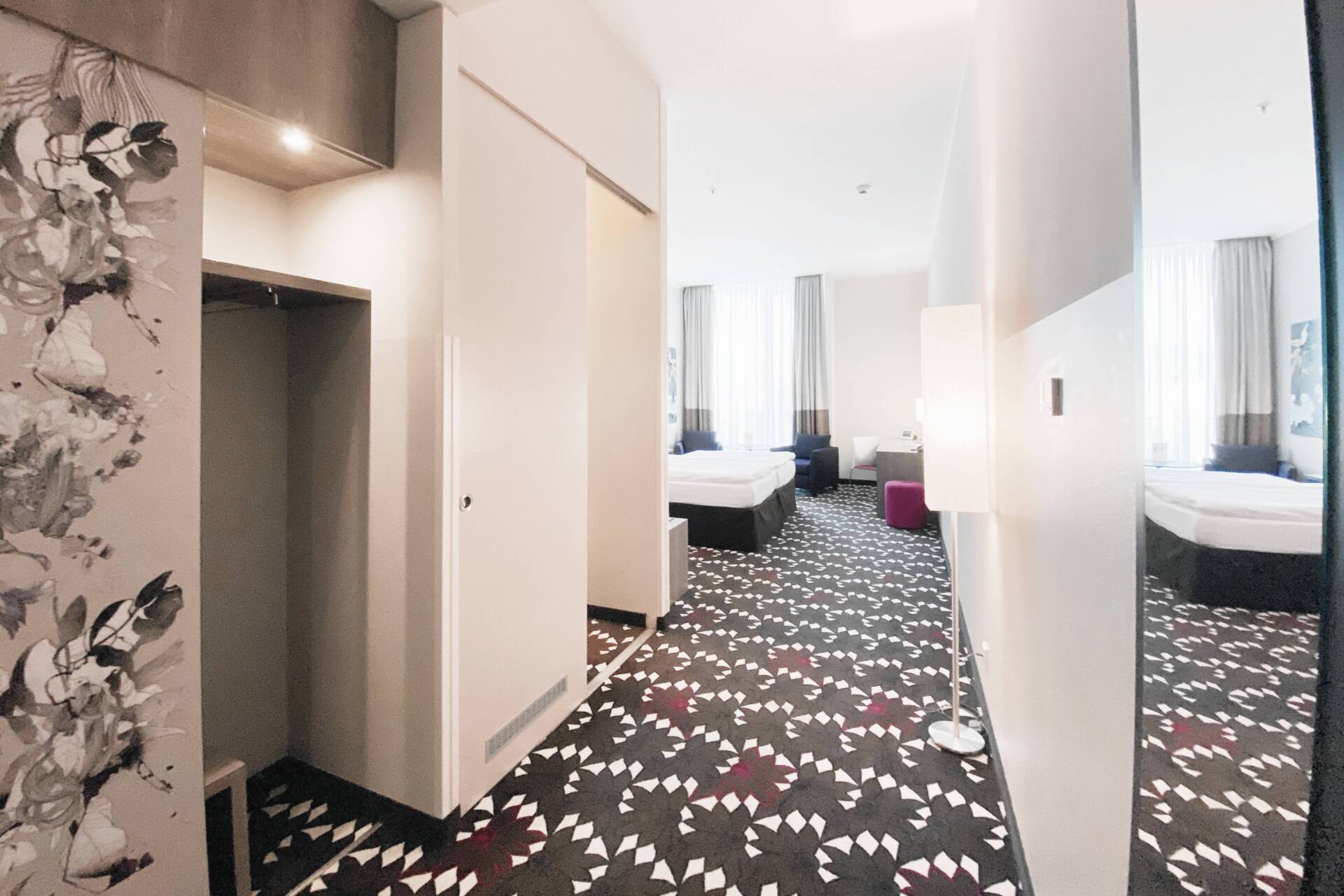 Helles Hotelzimmer mit zwei Betten, farbig gemustertem Teppichboden, Einbauschrank, Stehlampe und großem Wandspiegel.