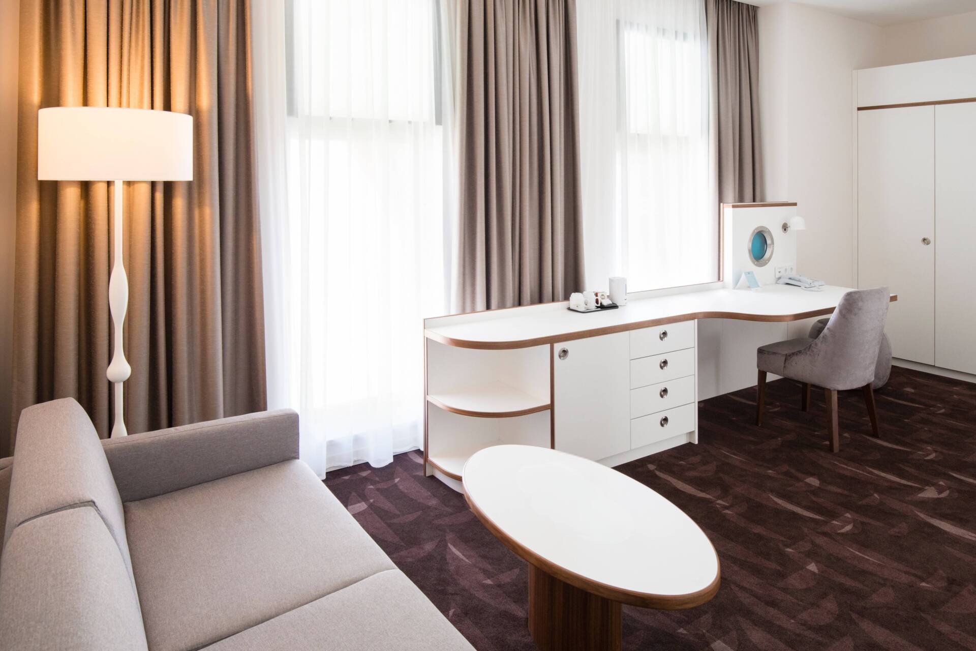 Modernly furnished hotel room with a desk, chair, sofa, coffee table, carpeted floor, and large windows with curtains.