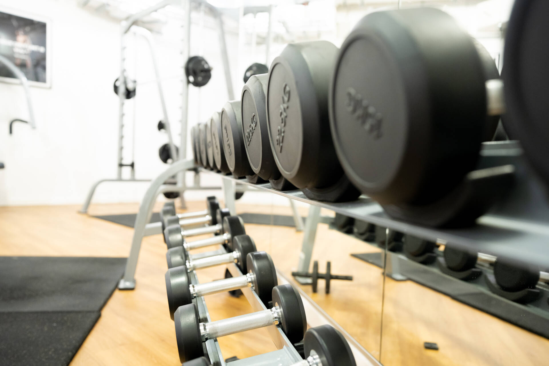 Reihe schwarzer Hanteln auf einem Gestell in einem hellen Fitnessraum mit Spiegelwand, Holzfußboden und weiteren Trainingsgeräten.