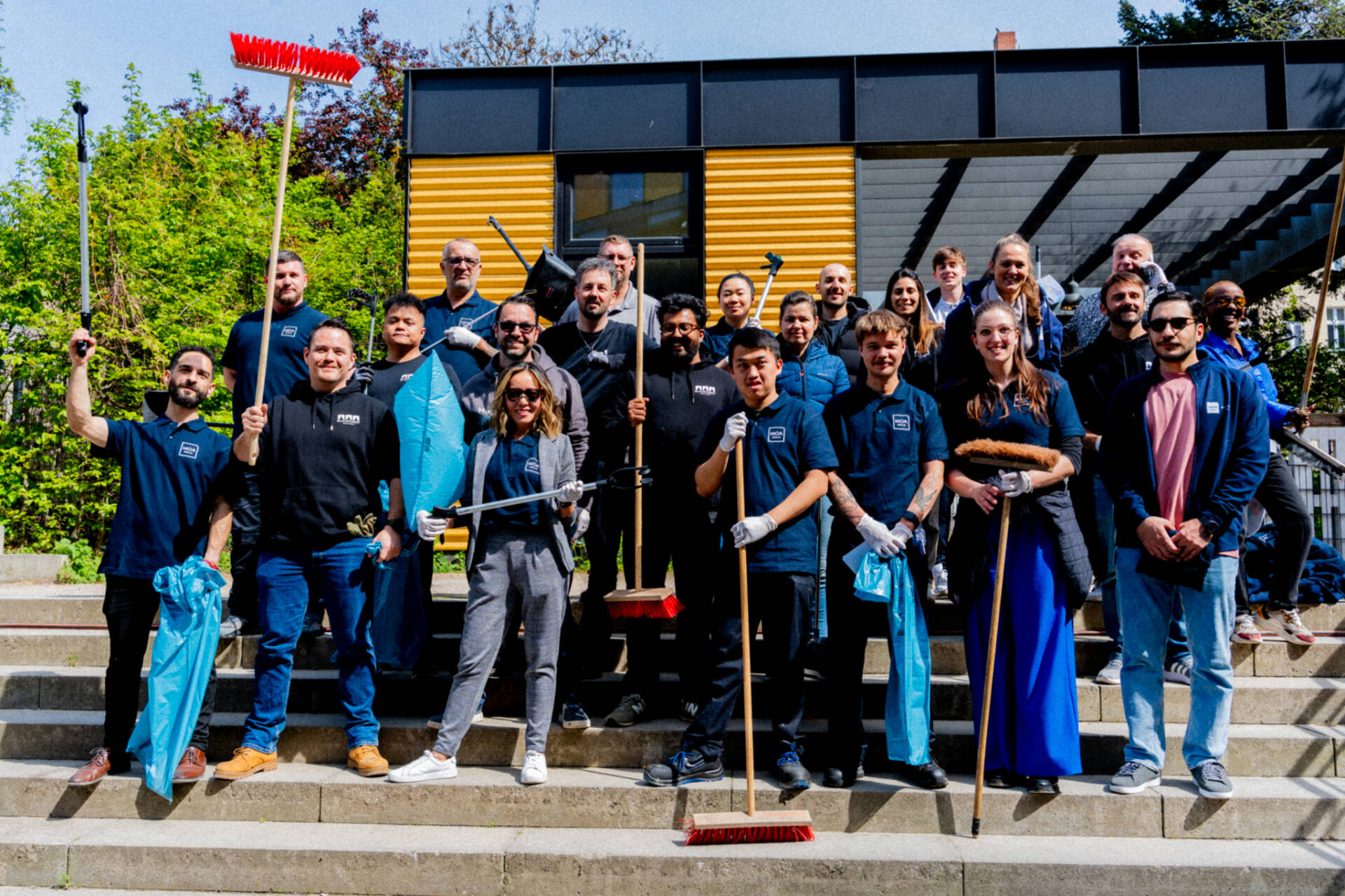 A group of people stands with brooms, litter pickers, and garbage bags on the steps in front of a modern building.