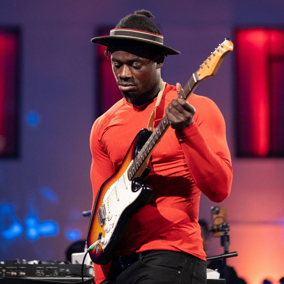 A musician wearing a hat plays an electric guitar in front of a brightly lit stage.