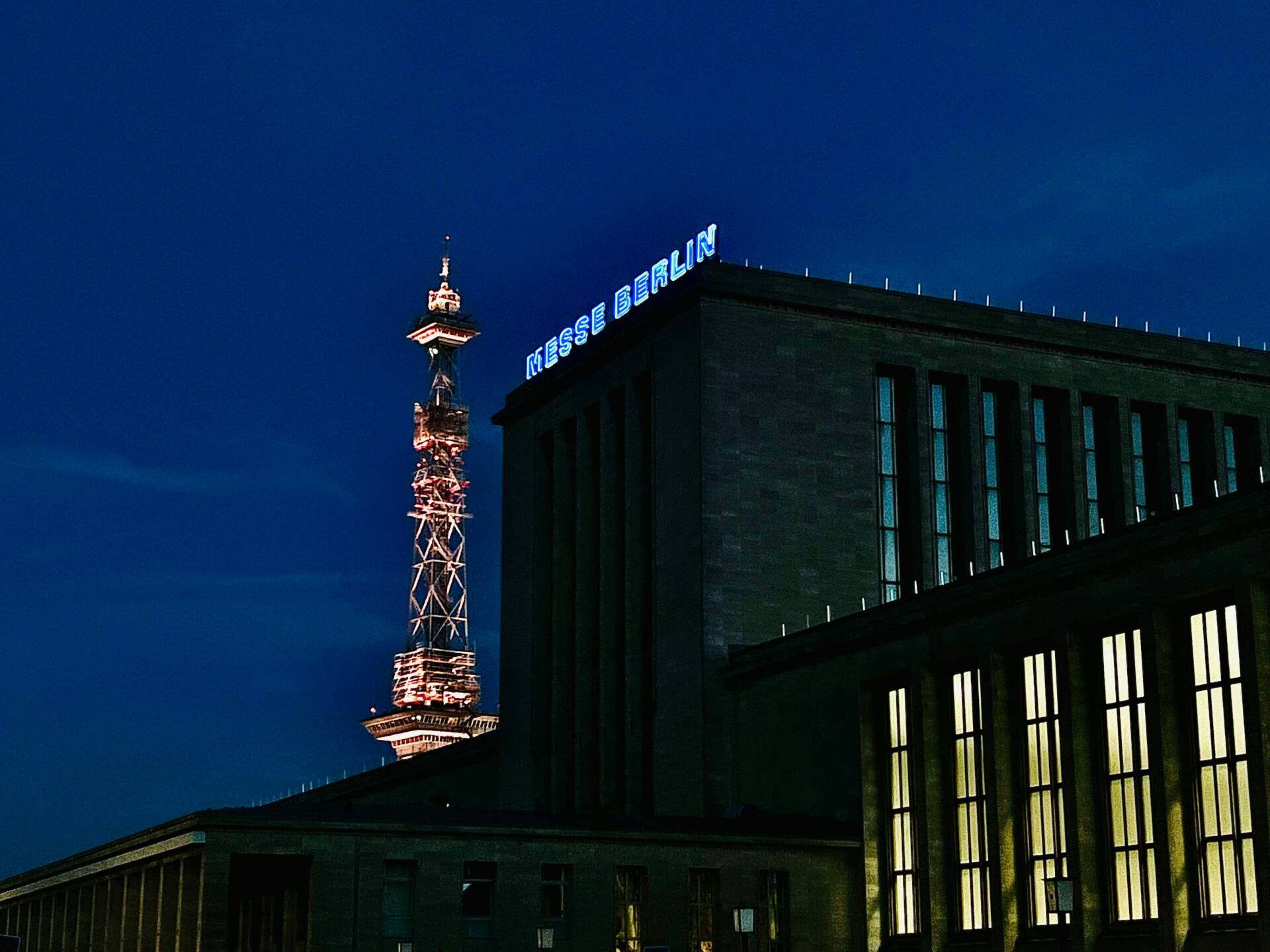 An illuminated building with the lettering „Messe Berlin“ and a transmission tower against a dark blue night sky.