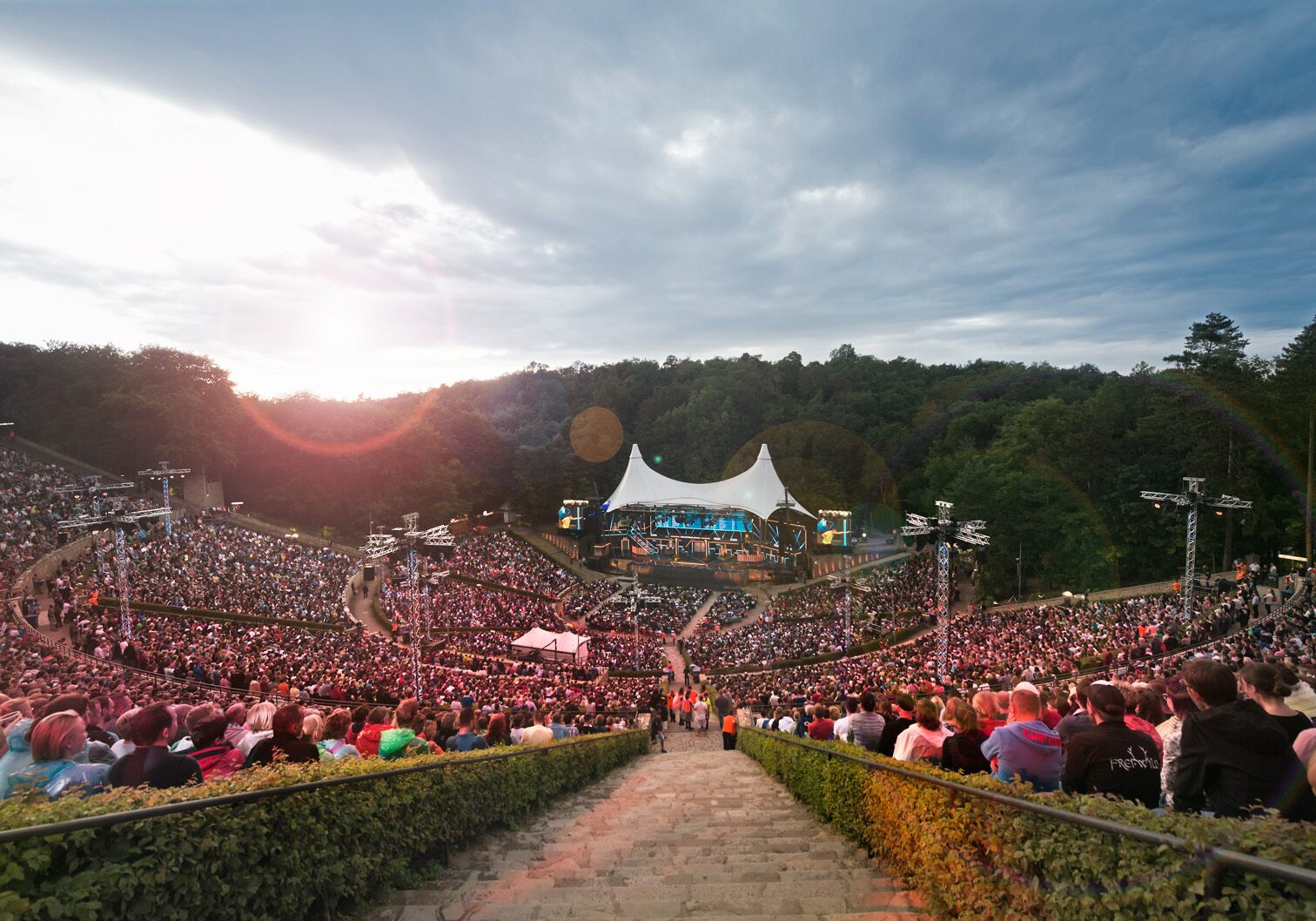 View of a large open-air event with a dense crowd, a stage and covered stands in a wooded landscape.
