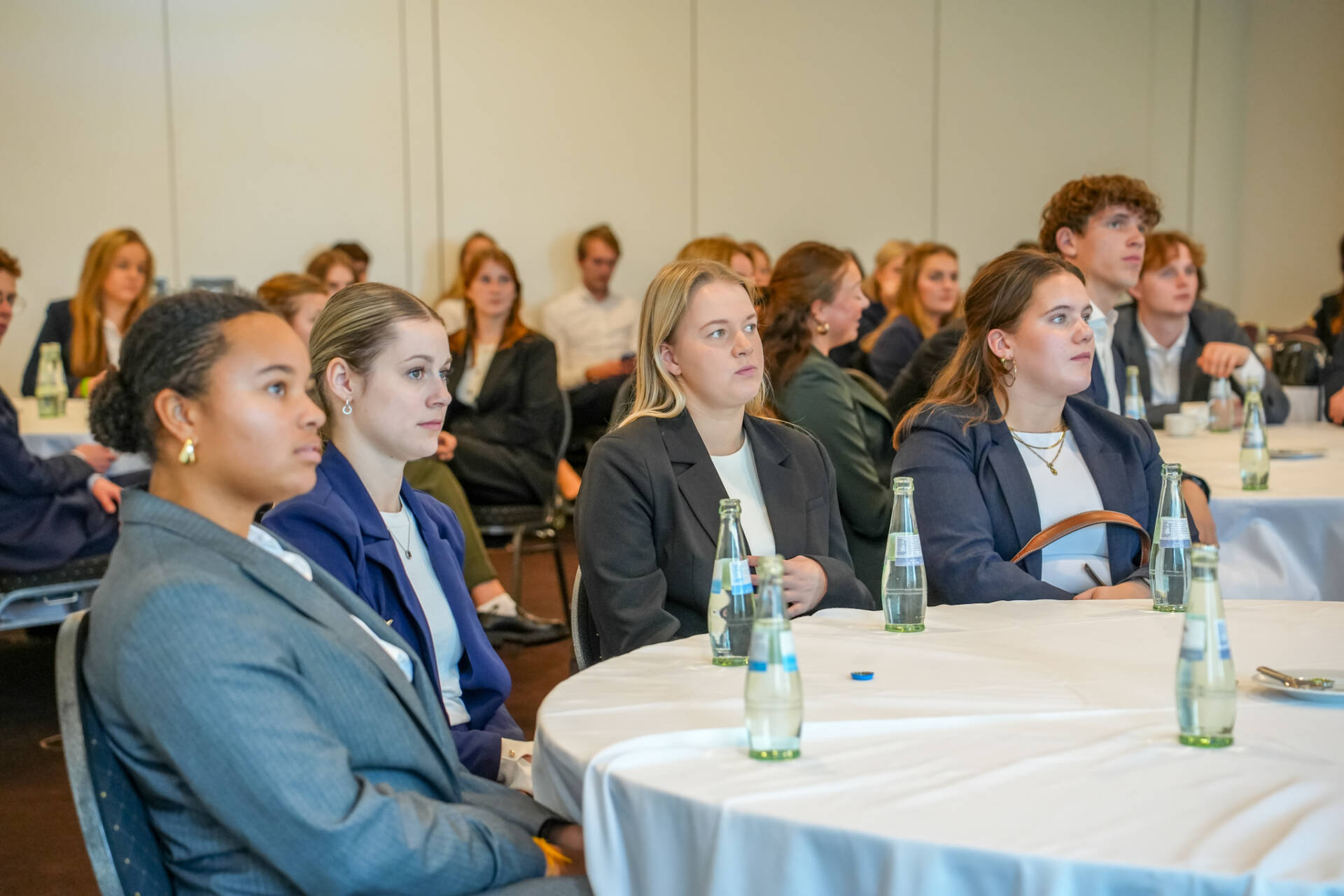 Mehrere Personen sitzen an runden Tischen in einem hellen Veranstaltungsraum und richten den Blick auf einen Bereich außerhalb des Bildes.