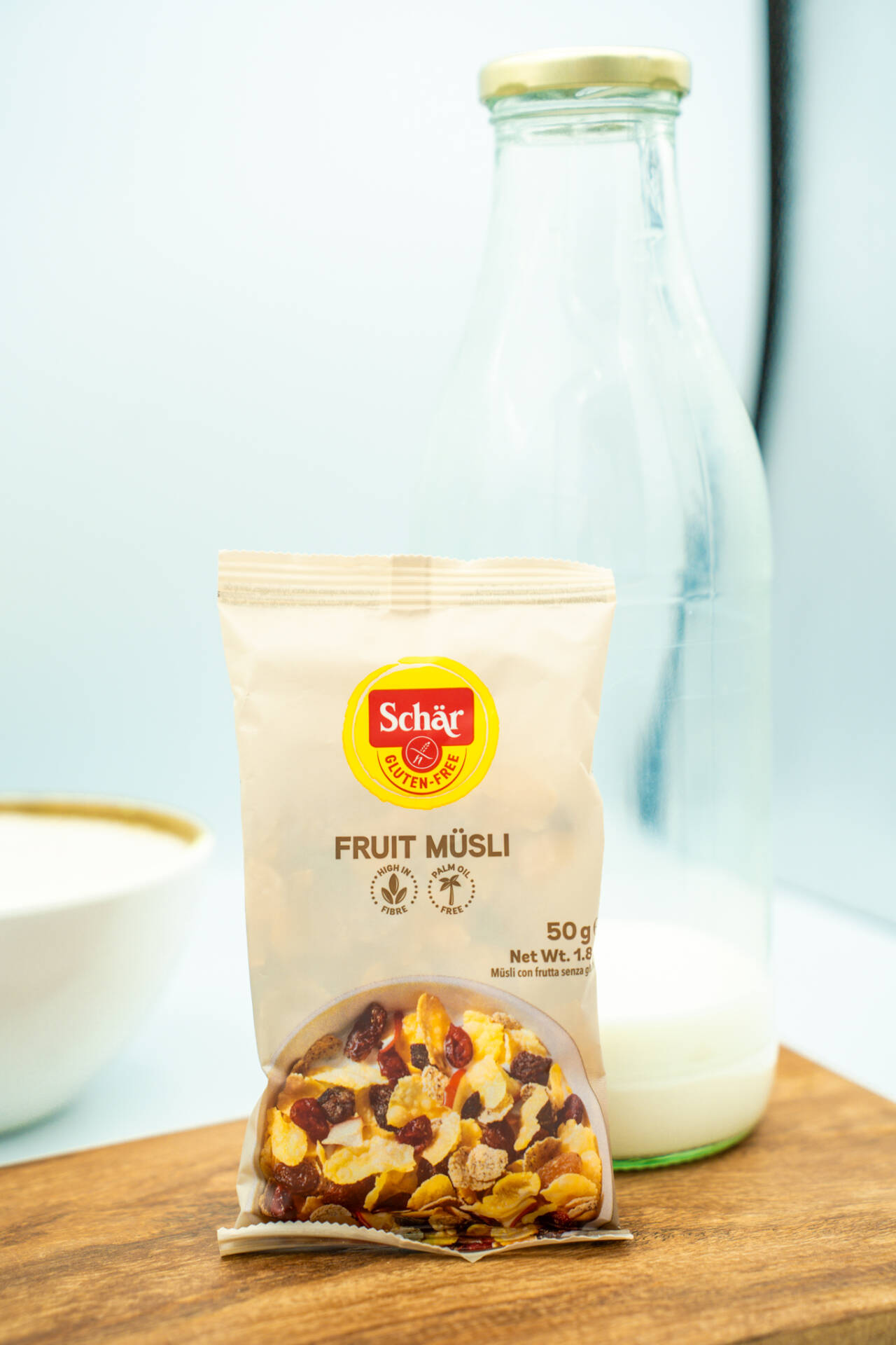 Eine Packung Schär Fruit Müsli mit 50 Gramm steht auf einem Holzbrett vor einer Glasflasche mit Milch.