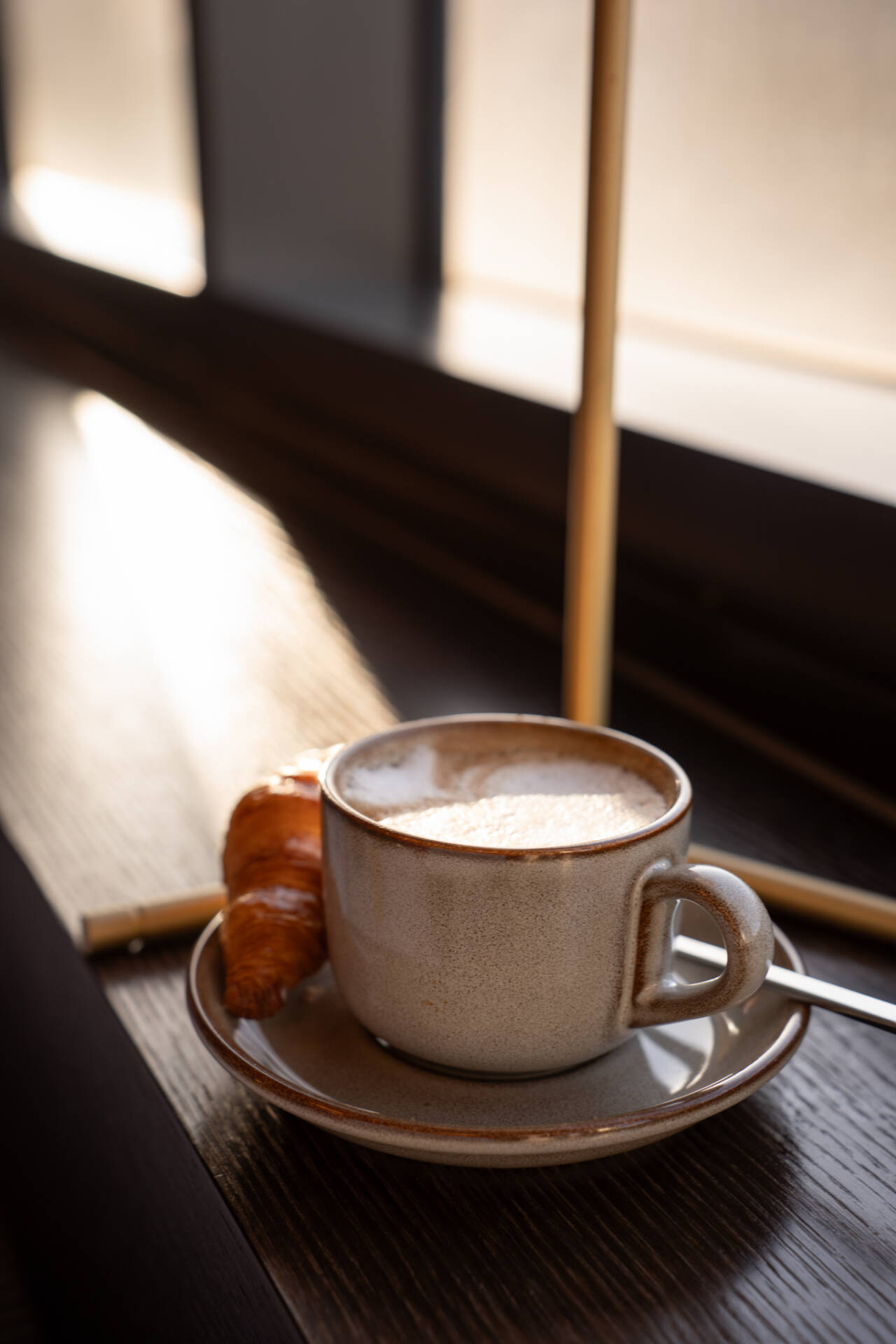 Eine Tasse Kaffee mit Milchschaum und ein Croissant stehen auf einem Unterteller an einem Tisch neben einem Fenster.