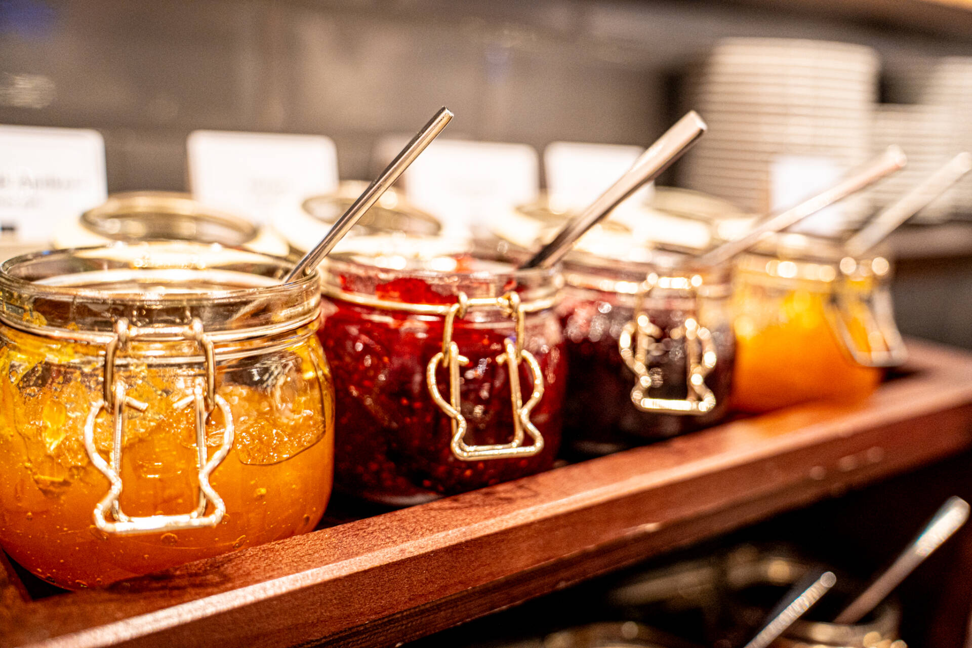 Mehrere Gläser mit verschiedenen Marmeladen und Löffeln stehen auf einem hölzernen Tablett am Frühstücksbuffet.
