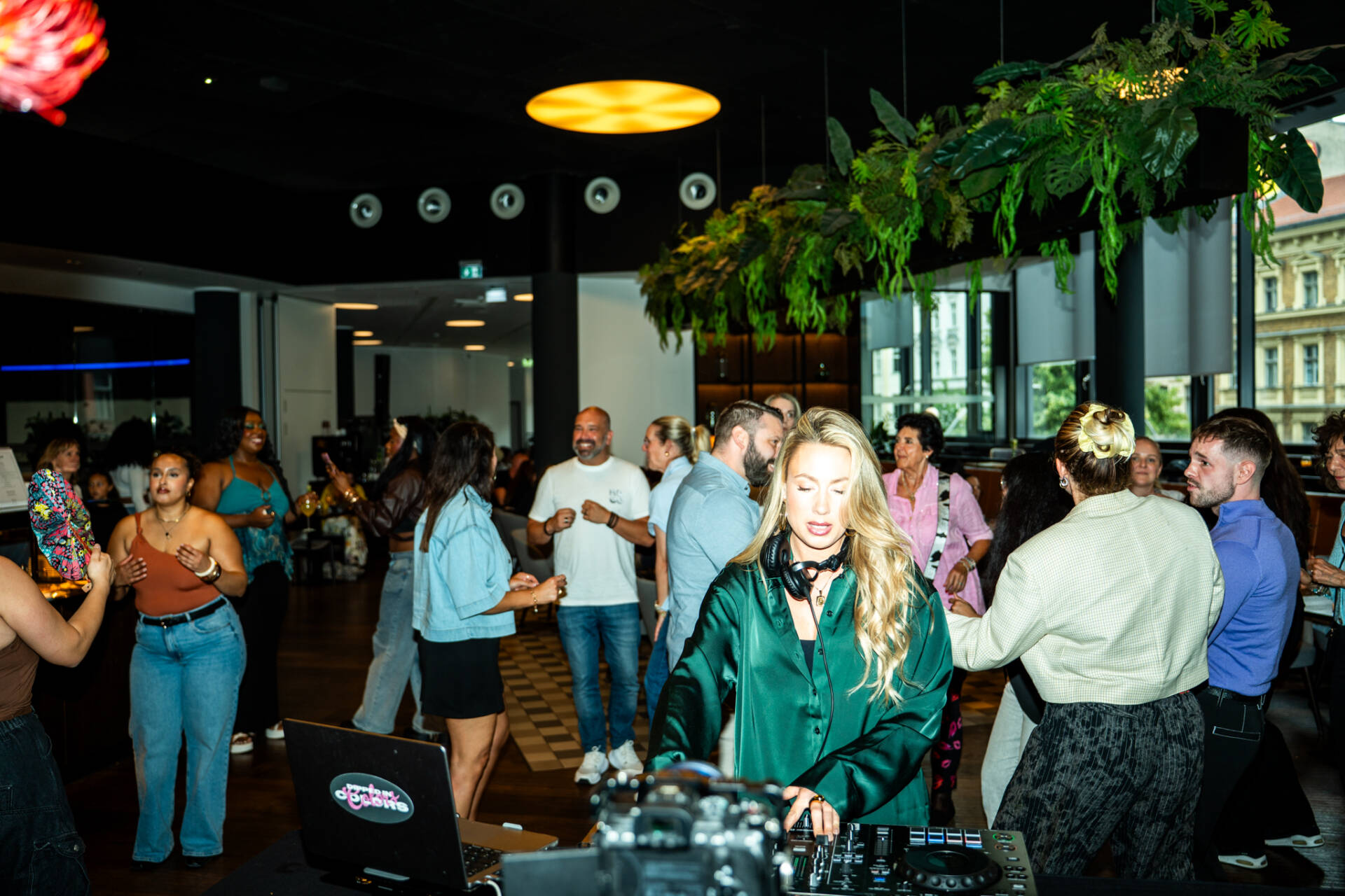 Menschen tanzen und unterhalten sich in einer Hotelbar, während eine DJane an einem Mischpult Musik auflegt.