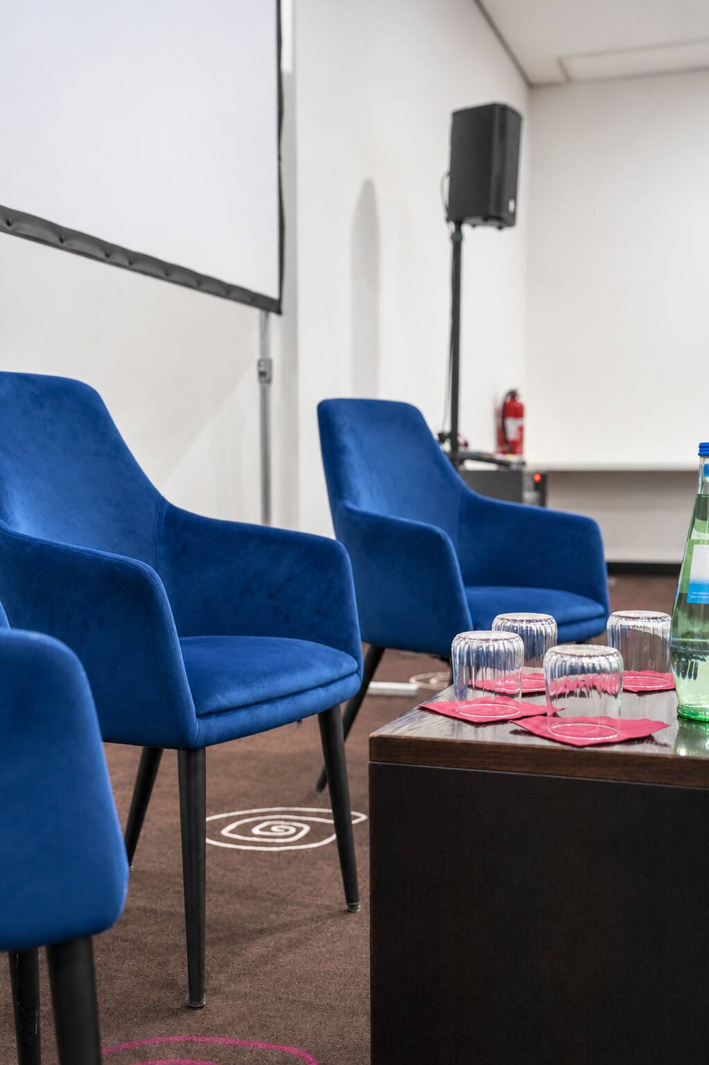 Blaue Sessel stehen in einem Konferenzraum neben einem Tisch mit Wassergläsern, pinken Untersetzern und einer grünen Flasche.