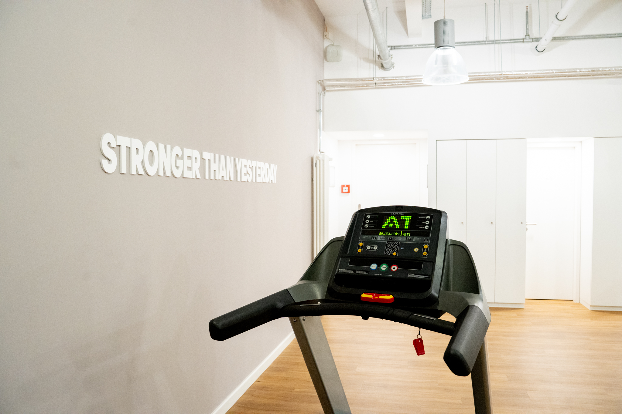 A treadmill stands in a bright fitness room with a wooden floor and the words "STRONGER THAN YESTERDAY" on the wall.