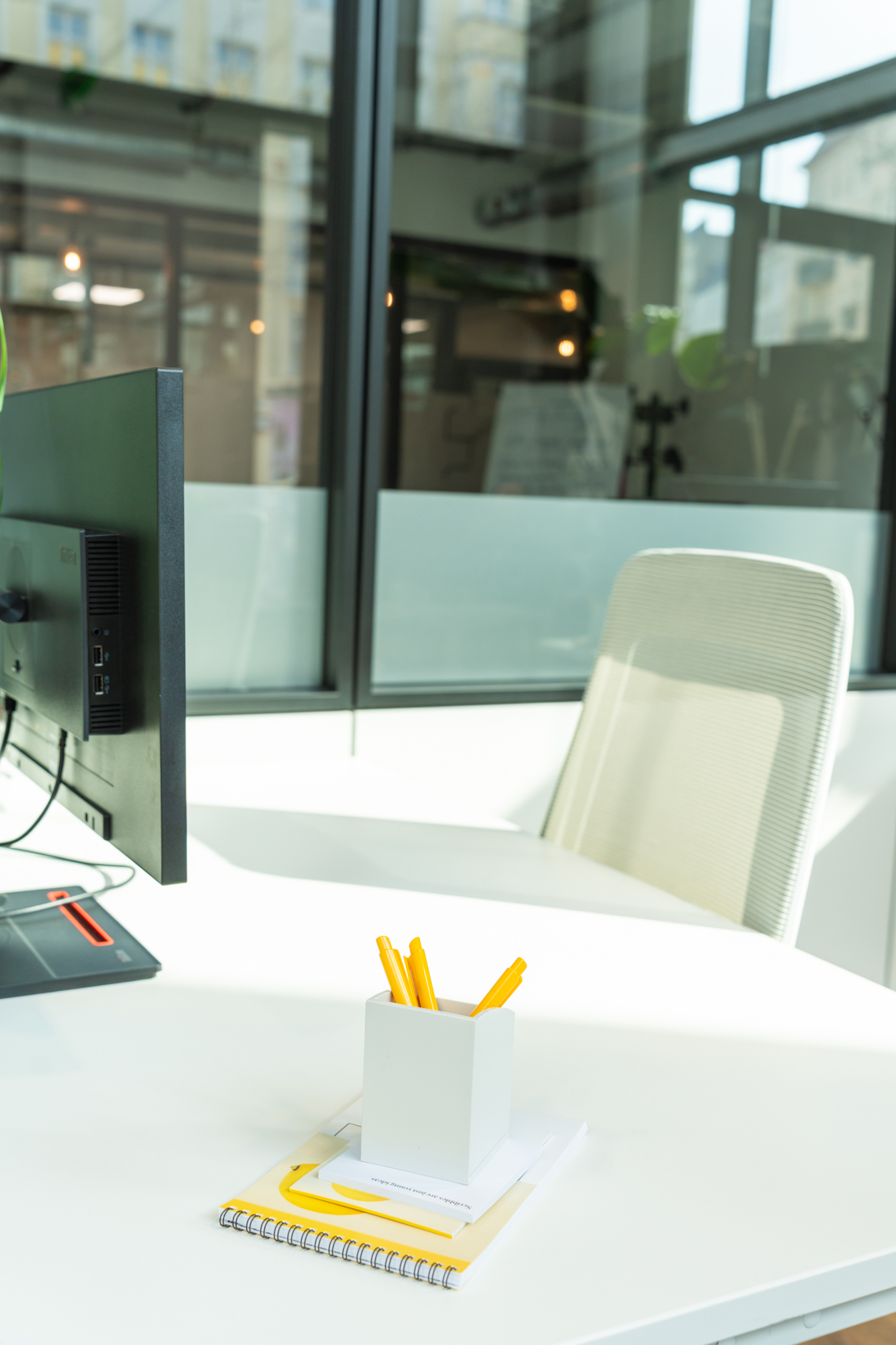 A bright workspace with a monitor, white office chair, pen holder and yellow notebook in front of large windows.