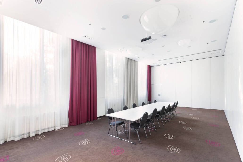 Ein heller Konferenzraum mit Tischen in Reihenanordnung, Stühlen, großen Fenstern und bodentiefen Vorhängen für Tagungen und Meetings.