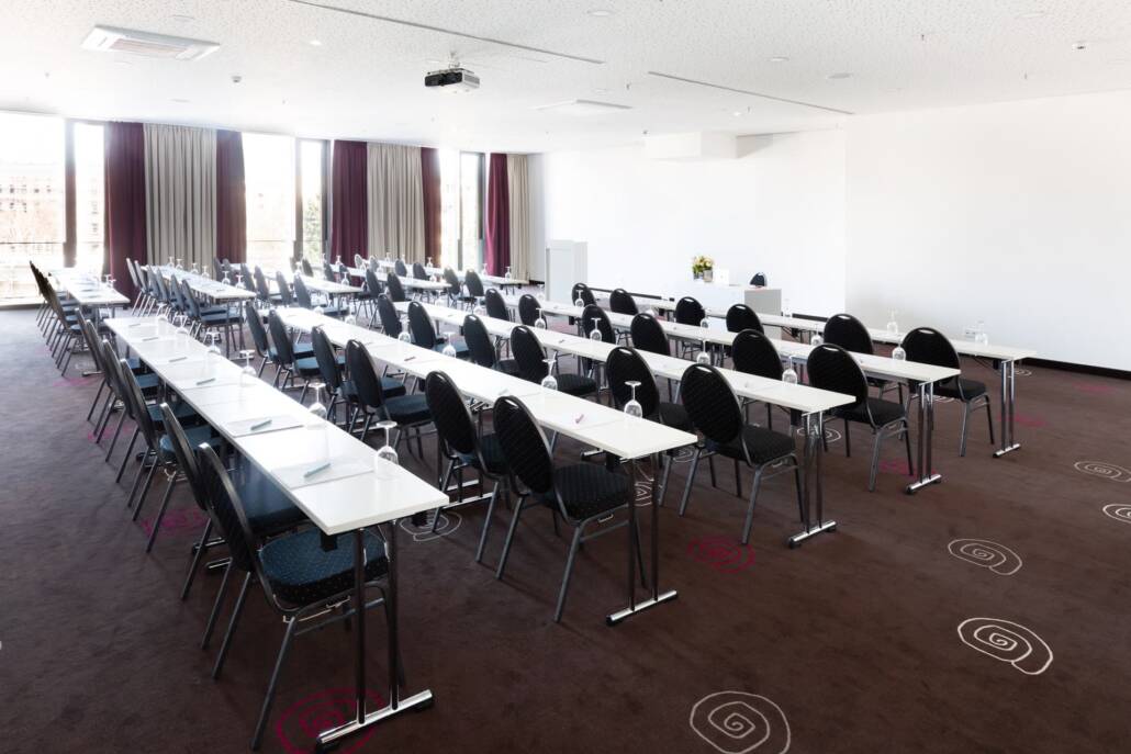 MOA_Meet_03_05_parlamentarische Bestuhlung_02-2 Ein heller Seminarraum mit einer parlamentarischen Reihenbestuhlung, Tischen, Stühlen, einer Leinwand und Fenstern.