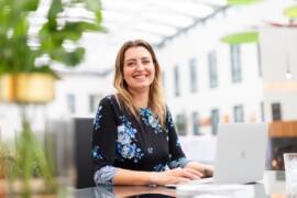 A receptionist with long hair wearing a black dress with a blue and white floral pattern sits at a table with a laptop in a bright, modern hotel in central Berlin.