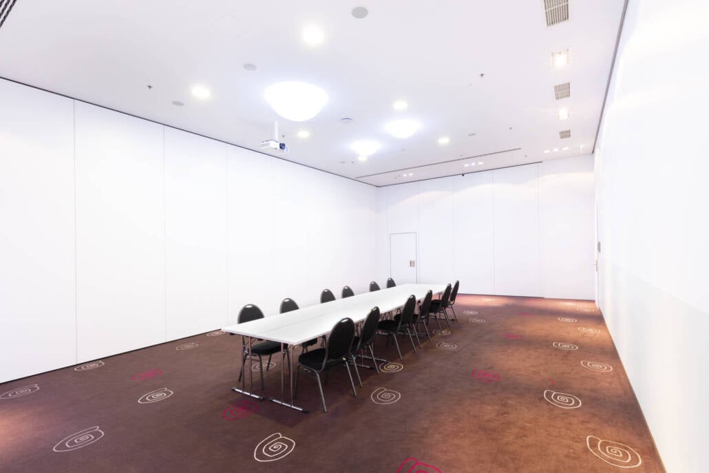 Ein großzügiger Veranstaltungsraum mit Teppichboden, Projektor, hoher Decke und Vorhängen für flexible Eventformate.