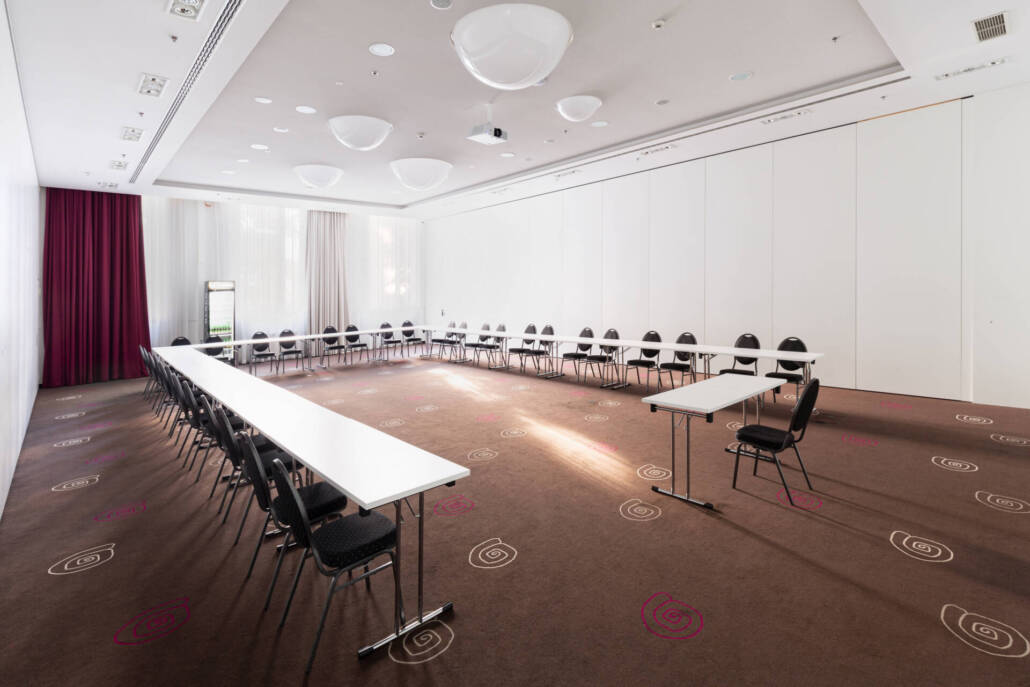 Ein moderner Konferenzraum mit einer U-Bestuhlung, Teppichboden, Deckenbeleuchtung und Tageslicht für Vorträge und Präsentationen.