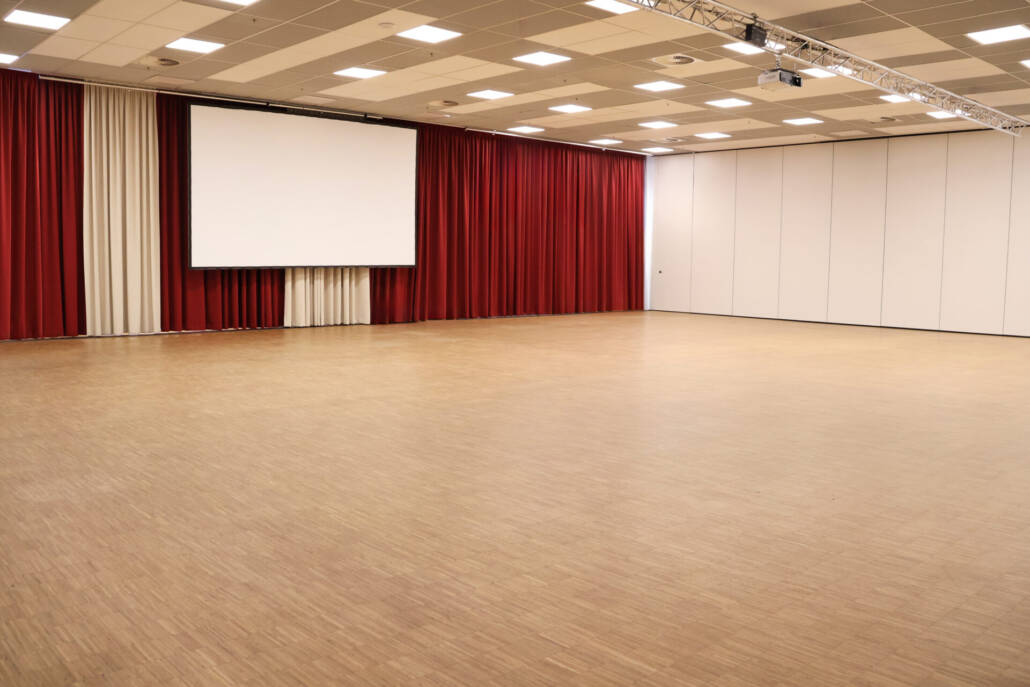 Ein großzügiger Eventraum mit einer Leinwand, einem roten Vorhang, Holzfußboden und einer technischen Infrastruktur für Veranstaltungen unterschiedlicher Formate.