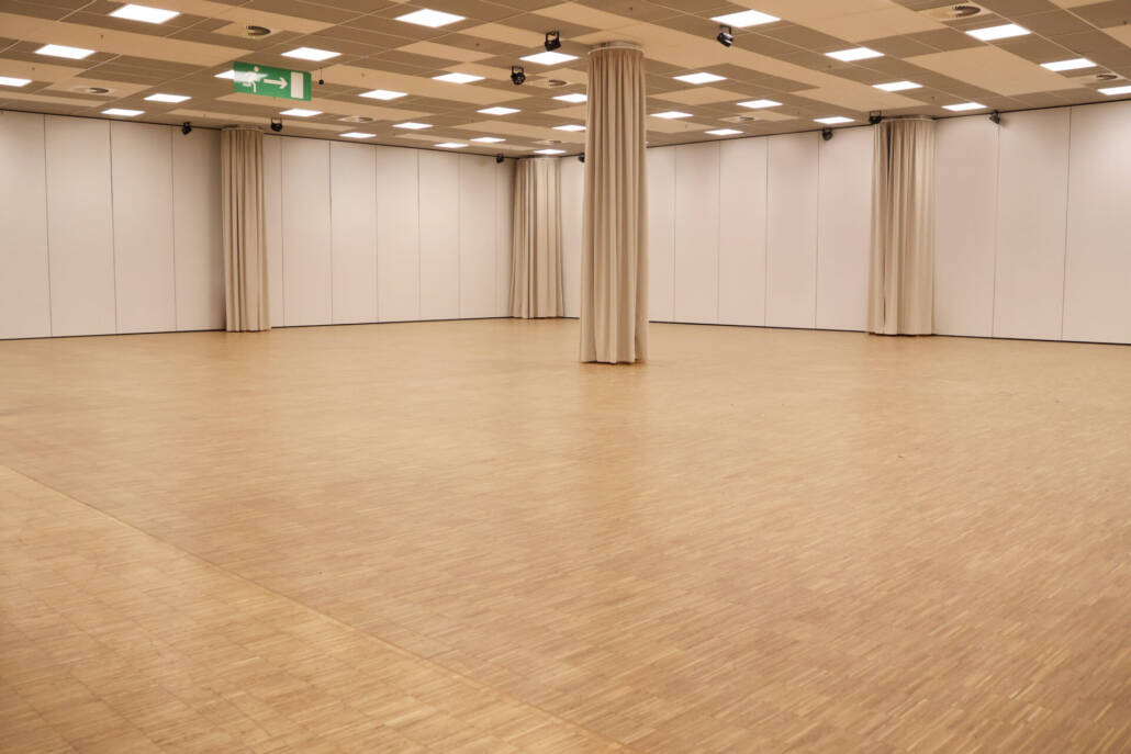 Ein großer, leerer Veranstaltungsraum mit einer hellen Deckenbeleuchtung, Säulen, einem Holzfußboden und flexiblen Trennwänden.