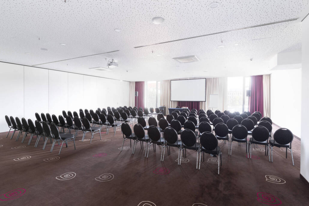 Ein professioneller Veranstaltungsraum mit einer Reihenbestuhlung, Bühne, Leinwand und Lichttechnik für Konferenzen und Events.