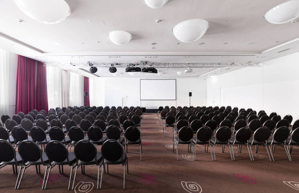 Ein professioneller Veranstaltungsraum mit einer Reihenbestuhlung, Bühne, Leinwand und Lichttechnik für Konferenzen und Events.