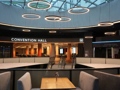 A modern convention hall with escalators, seating areas, round ceiling lighting and a view into a prepared event room.