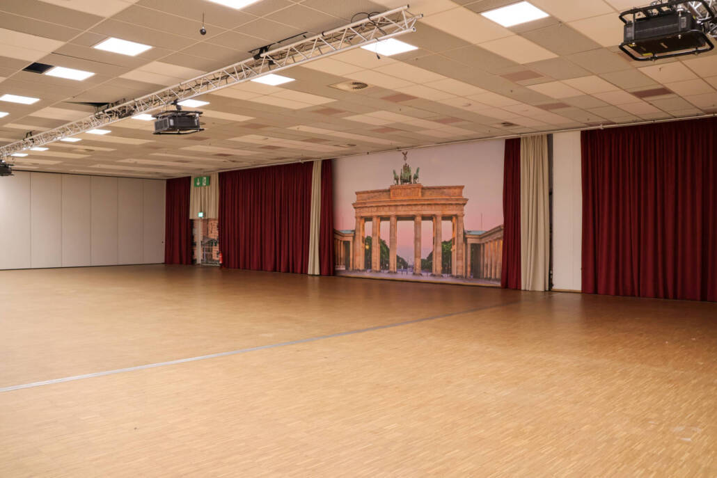 Ein großzügiger Veranstaltungsraum mit einem Holzfußboden, einem roten Vorhang, einer Leinwand und einem dekorativen Bühnenhintergrund für Events und Präsentationen.