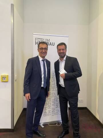 Zwei Männer in Anzügen stehen vor einem Banner mit der Aufschrift "FORUM HOCHBAU" in einem hellen Raum des Hotels MOA Berlin.