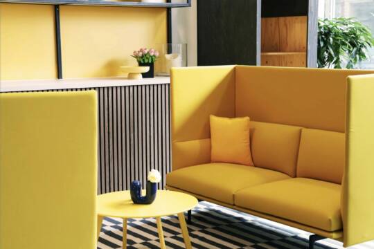 Yellow lounge area in the co-working space of the MOA Hotel Berlin with a modern yellow sofa, high side walls, a round yellow table and a dark blue vase with a white flower. In the background are decorative elements and a plant, and the floor is covered with a striped carpet.