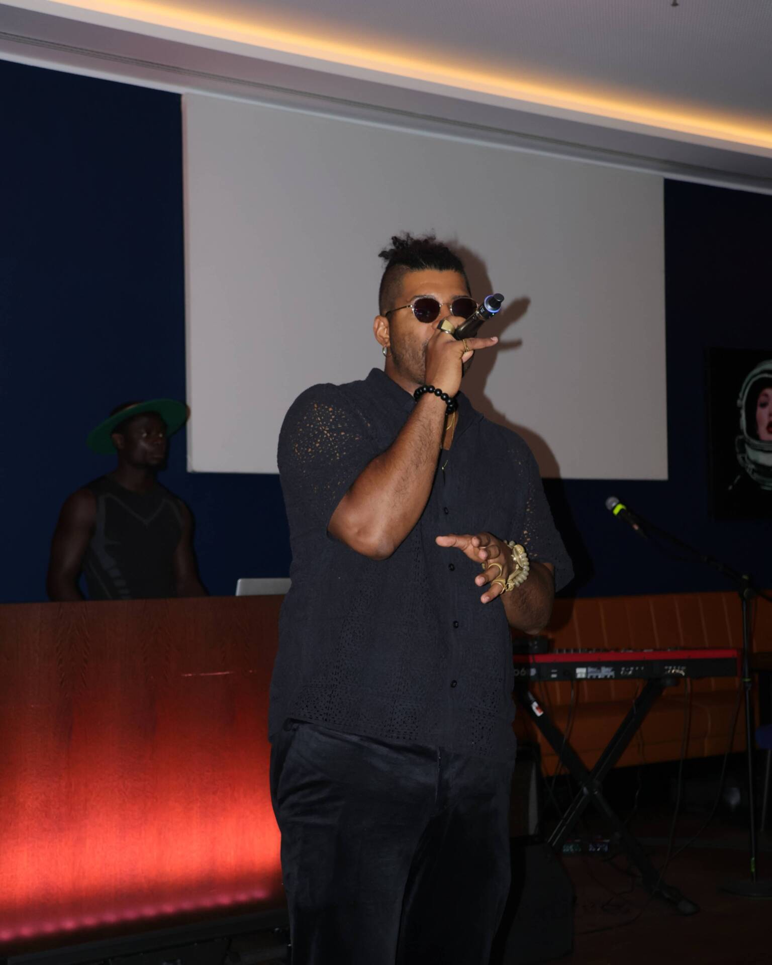 Ein Mann mit Mikrofon singt auf einer Bühne vor einem Keyboard und einem großen Bildschirm im Hintergrund.