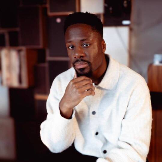 A seated man wears a light-coloured, long-sleeved jacket with buttons and holds one hand to his chin. Shelves with books and decorative items can be seen in the background.